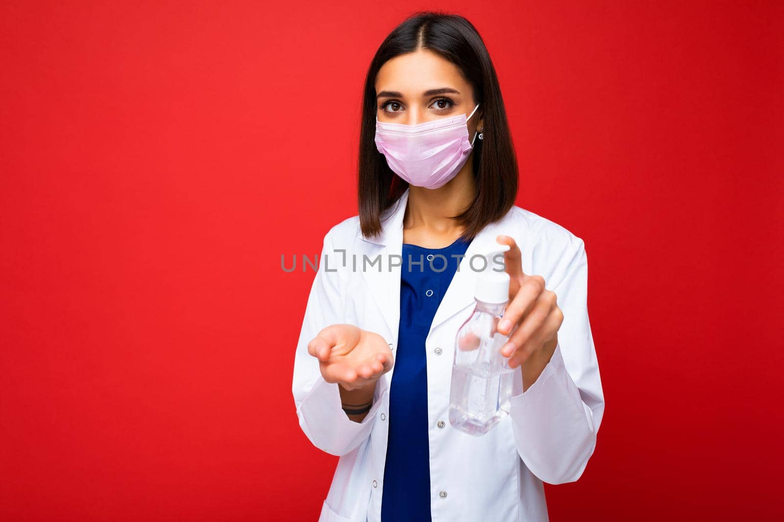 Young beautiful woman in a white doctors coat and a medical mask on her face splashes antiseptics on her hands to prevent infections isolated on a background.