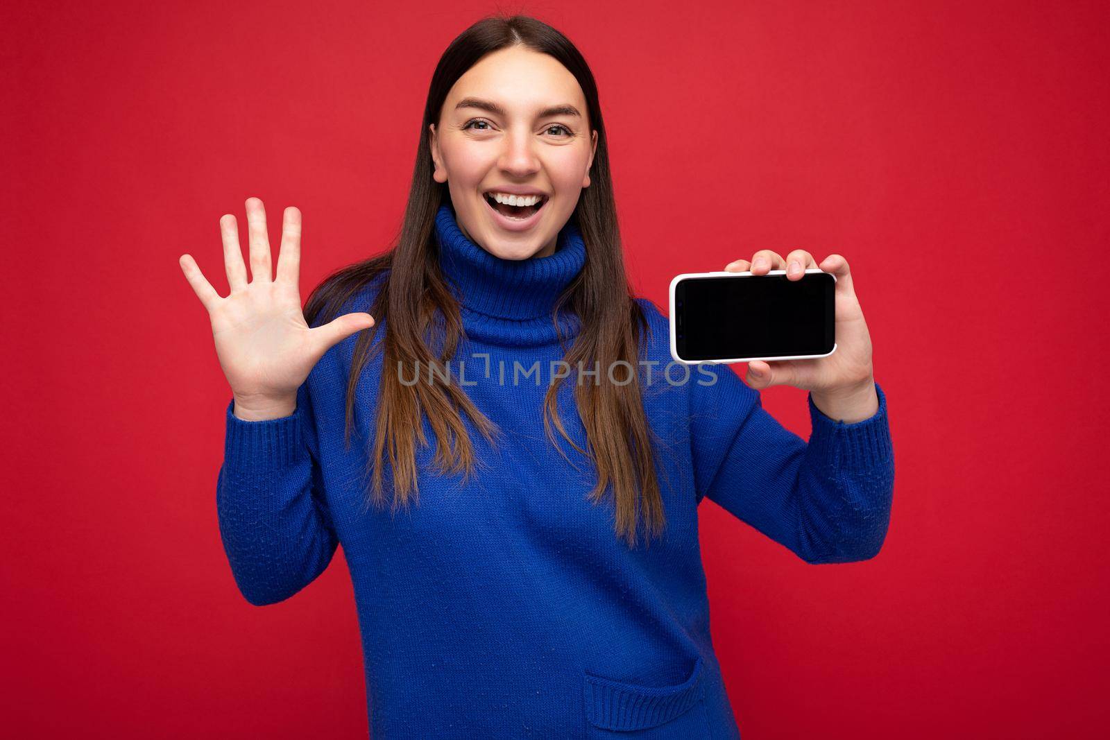 Closeup portrait photo shot of charming happy young brunette woman wearing casual blue sweater isolated over red background with empty space holding in hand mobile phone and showing smartphone with empty screen for mockup looking at camera and showing open palm and five fingers.
