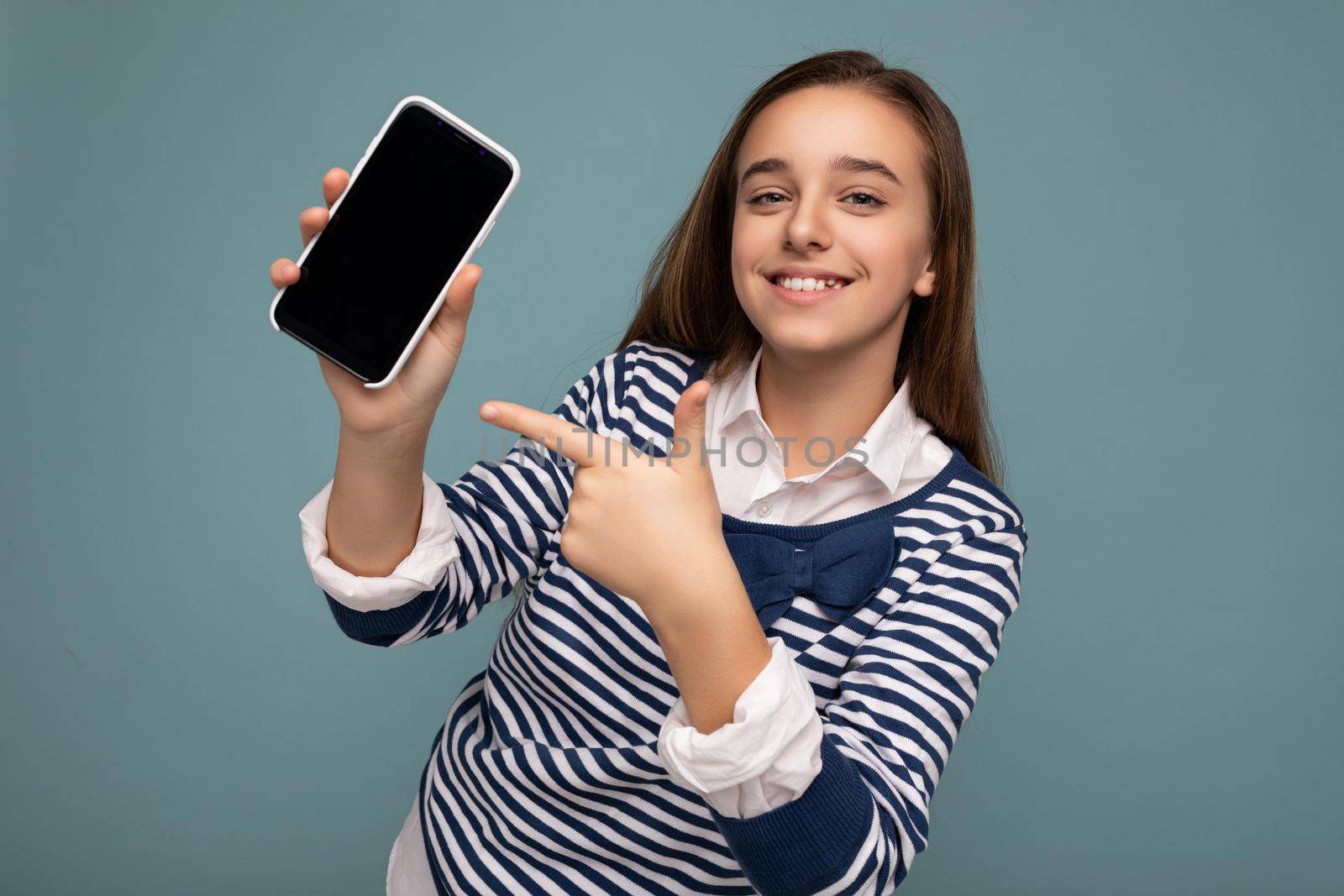 Photo of beautiful smiling girl good looking wearing casual stylish outfit standing isolated on background with copy space holding smartphone showing phone in hand with empty screen display for mockup pointing at gadjet looking at camera.