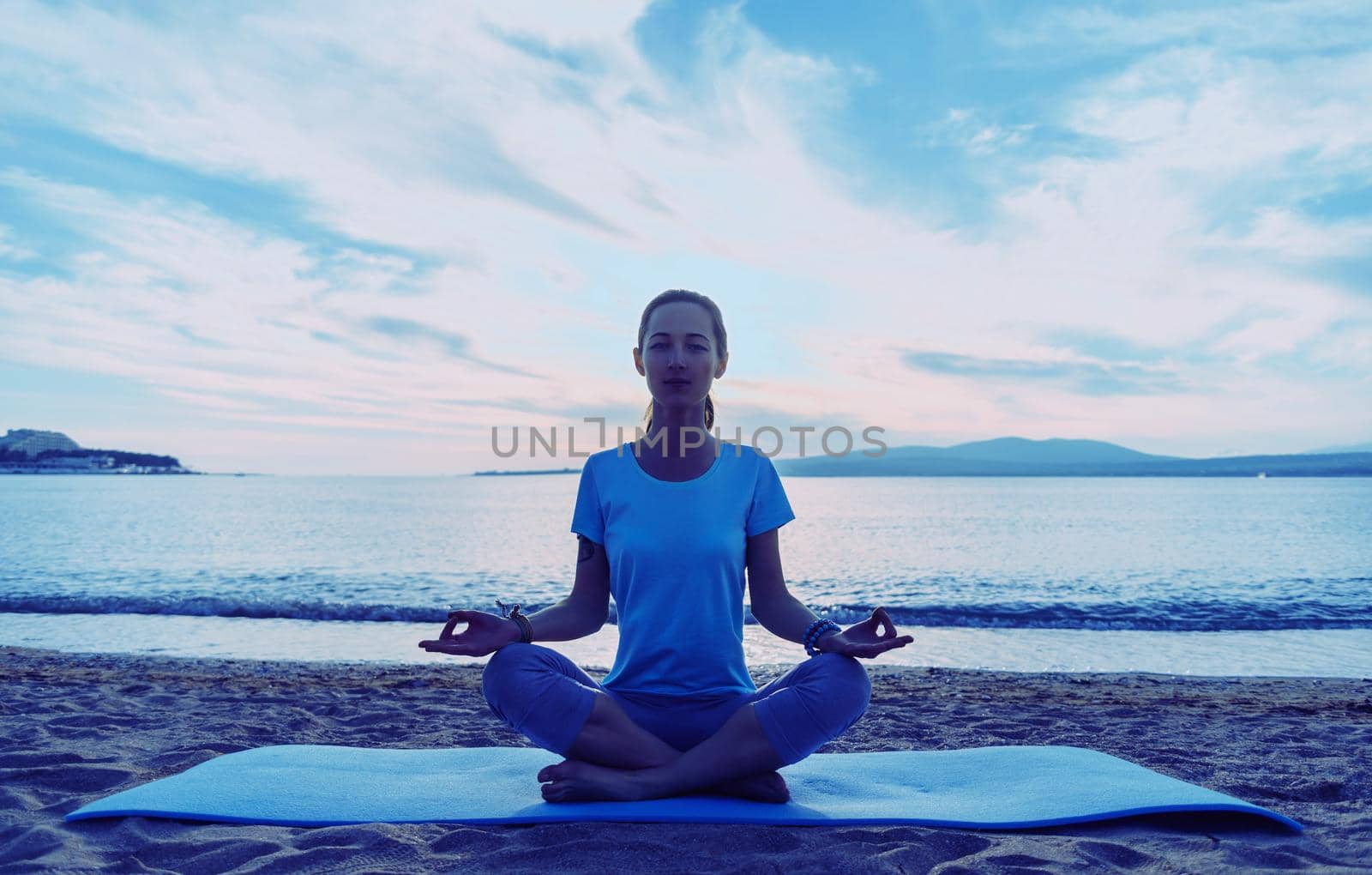 Young woman meditating in pose of lotus on sand beach near the sea in summer in the morning, front view