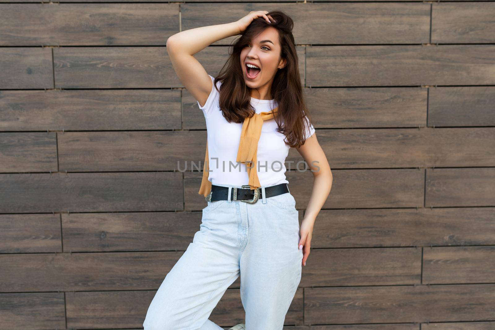 Portrait of successful smiling joyful happy young brunet woman wearing casual white t-shirt and jeans with yellow sweater poising near brown wall in the street and having fun.