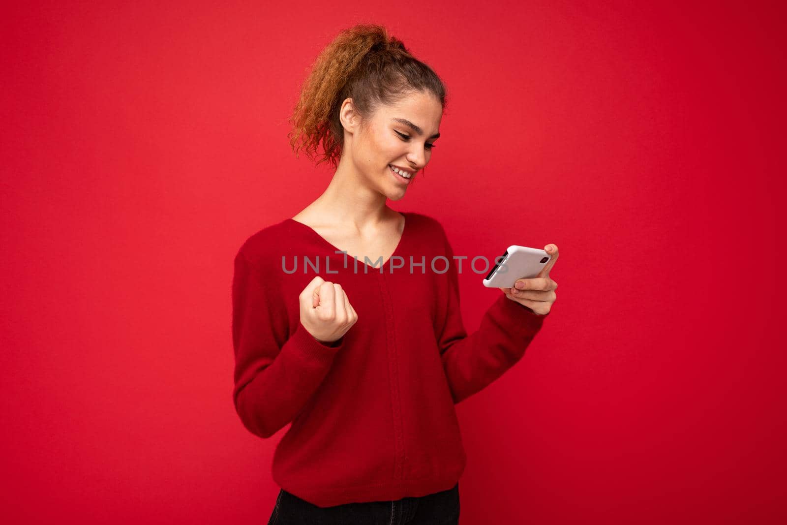 Cheerful woman standing isolated over red background, holding mobile phone, celebrating.