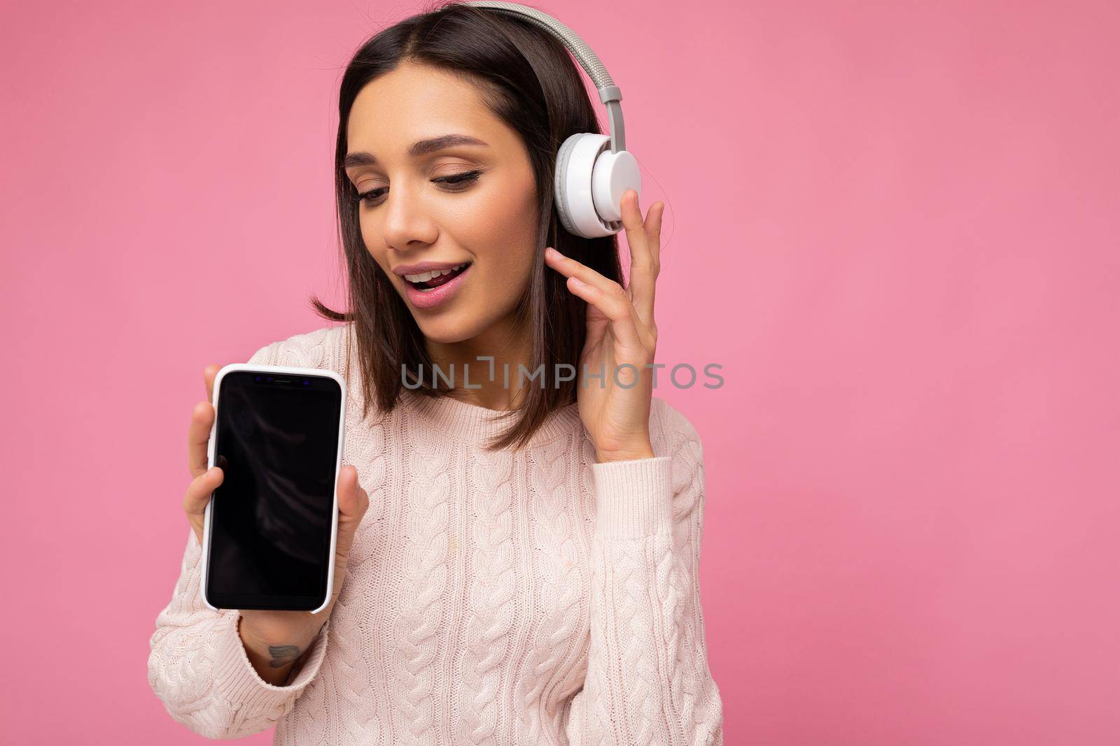 Closeup portrait of attractive positive cute young woman wearing stylish casual outfit isolated on colourful background wall holding and showing mobile phone with empty screen for cutout wearing white bluetooth headphones and having fun looking down.