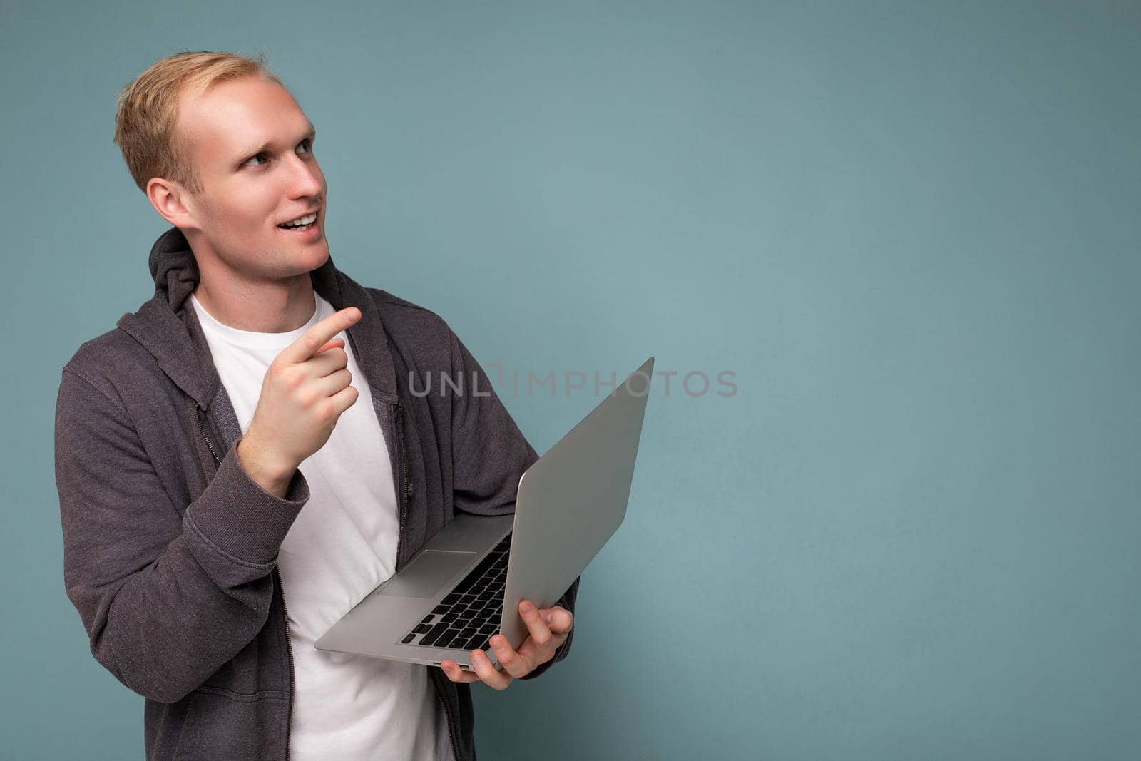 Side profile photo shot of handsome thoughtful blonde man holding computer laptop wearing white t-shirt and grey sweater looking up to the side thinking over isolated over blue background. Copy space