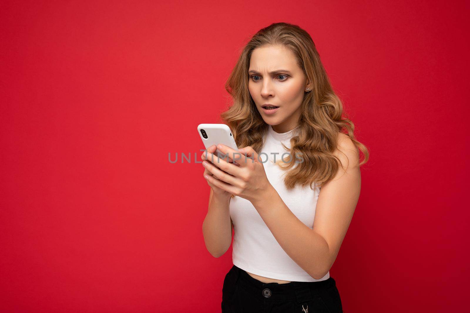 Amazed shocked beautiful young blonde woman wearing white t-shirt isolated over red background using smartphone and chatting via mobile phone looking at gadjet screen.