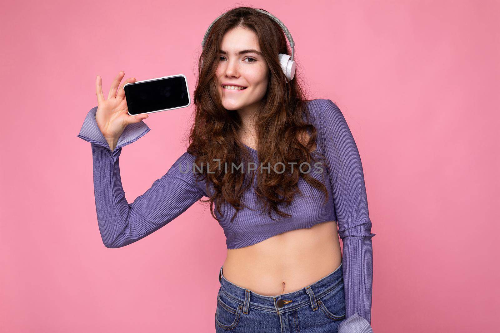 Beautiful happy smiling young woman wearing stylish casual outfit isolated on background wall holding and showing mobile phone with empty display for mockup wearing white bluetooth headphones listening to music and having fun looking at camera.