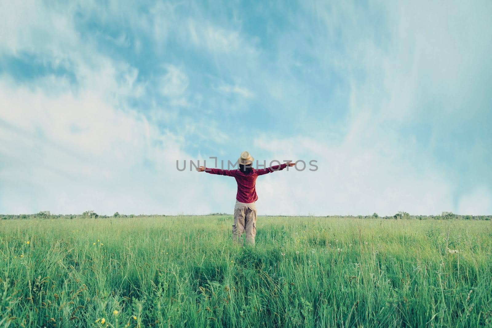 Young woman in straw hat standing with raised arms on meadow in summer. Concept of happiness and carefree