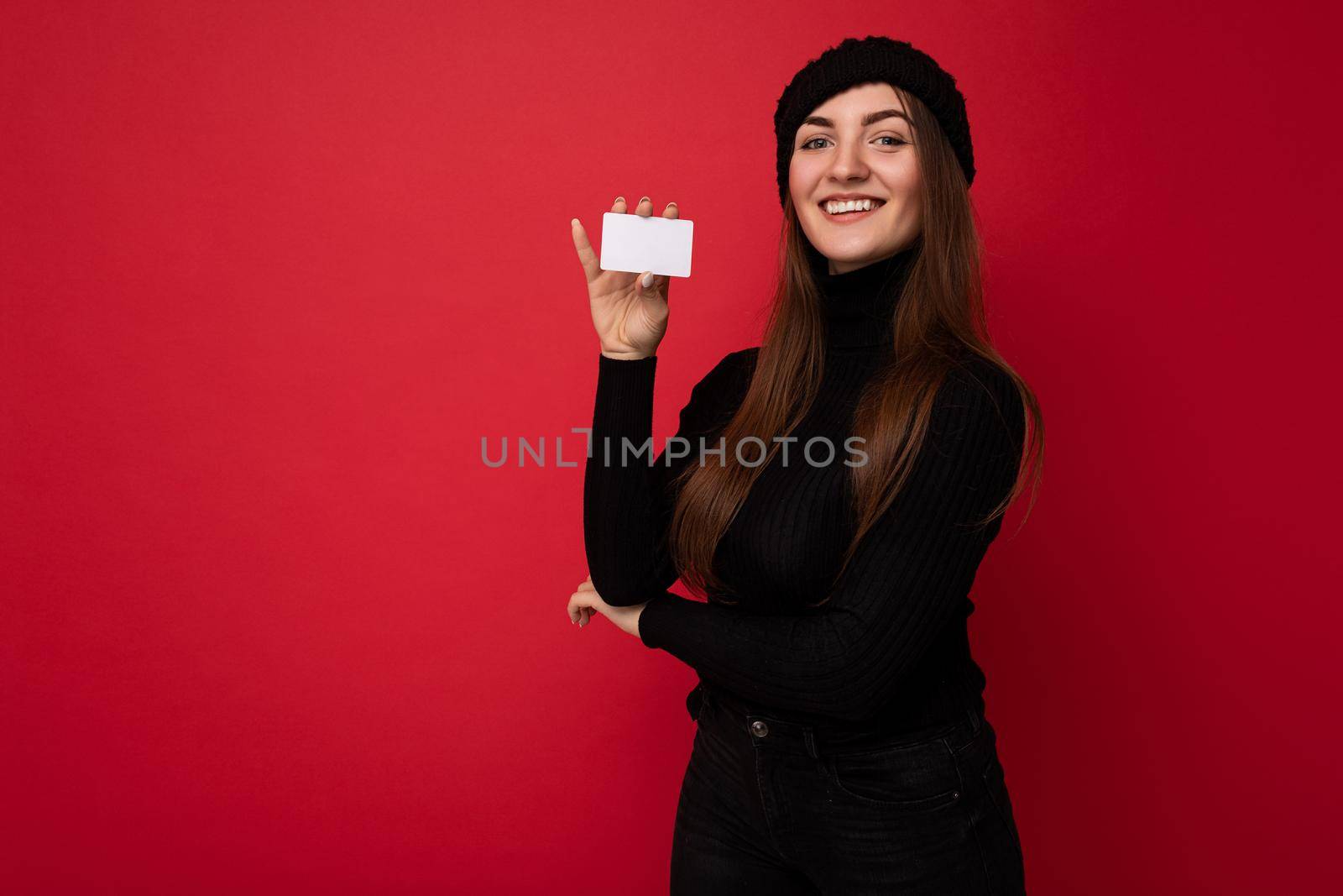 Photo of beautiful positive young brunette woman wearing black sweater and hat isolated on red background holding credit card looking at camera. copy space