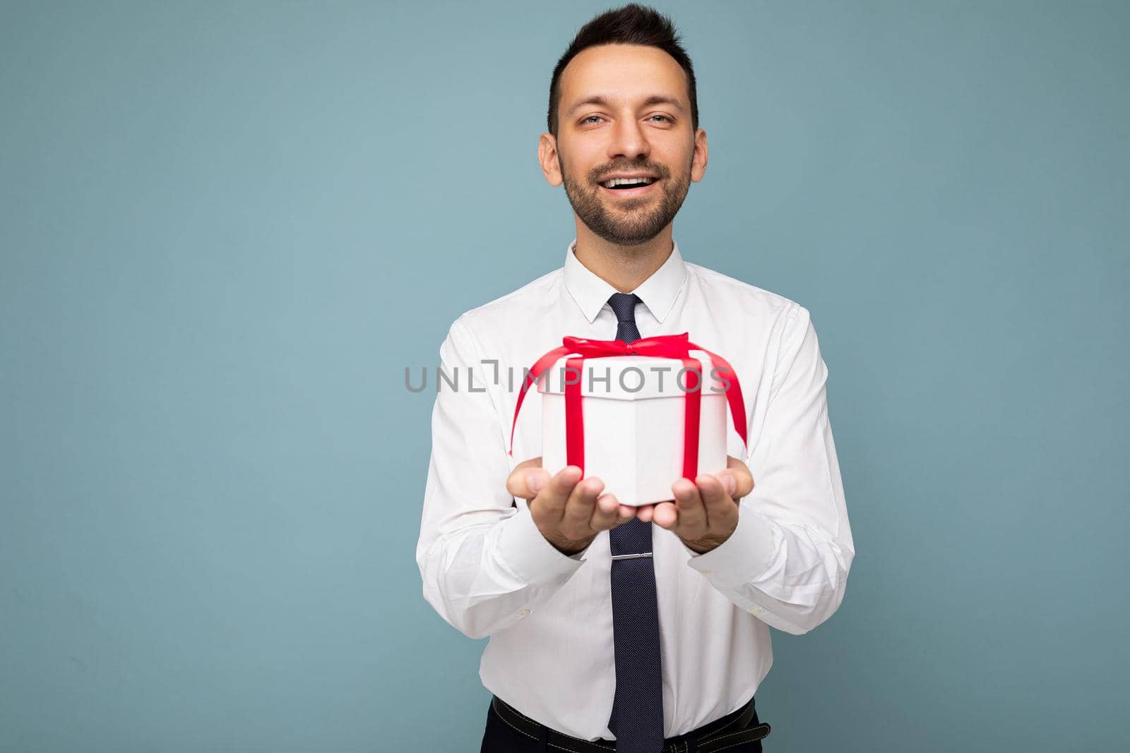 Photo shot of handsome positive brunette unshaven young man isolated over blue background wall wearing white shirt holding white gift box with red ribbon and looking at camera.