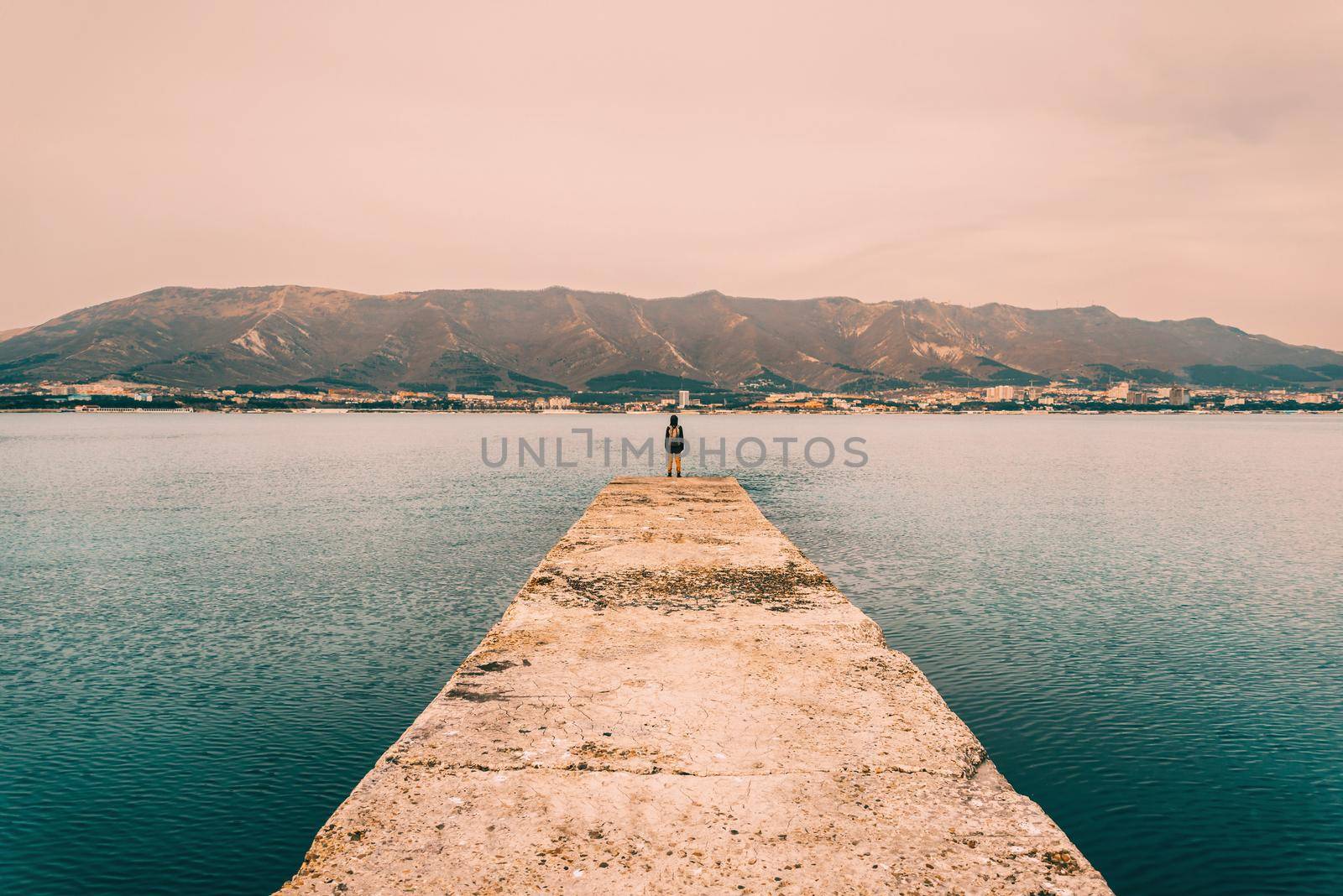 Hiker woman with backpack standing on pier at bay in front of mountains. Image with color effect, green toning