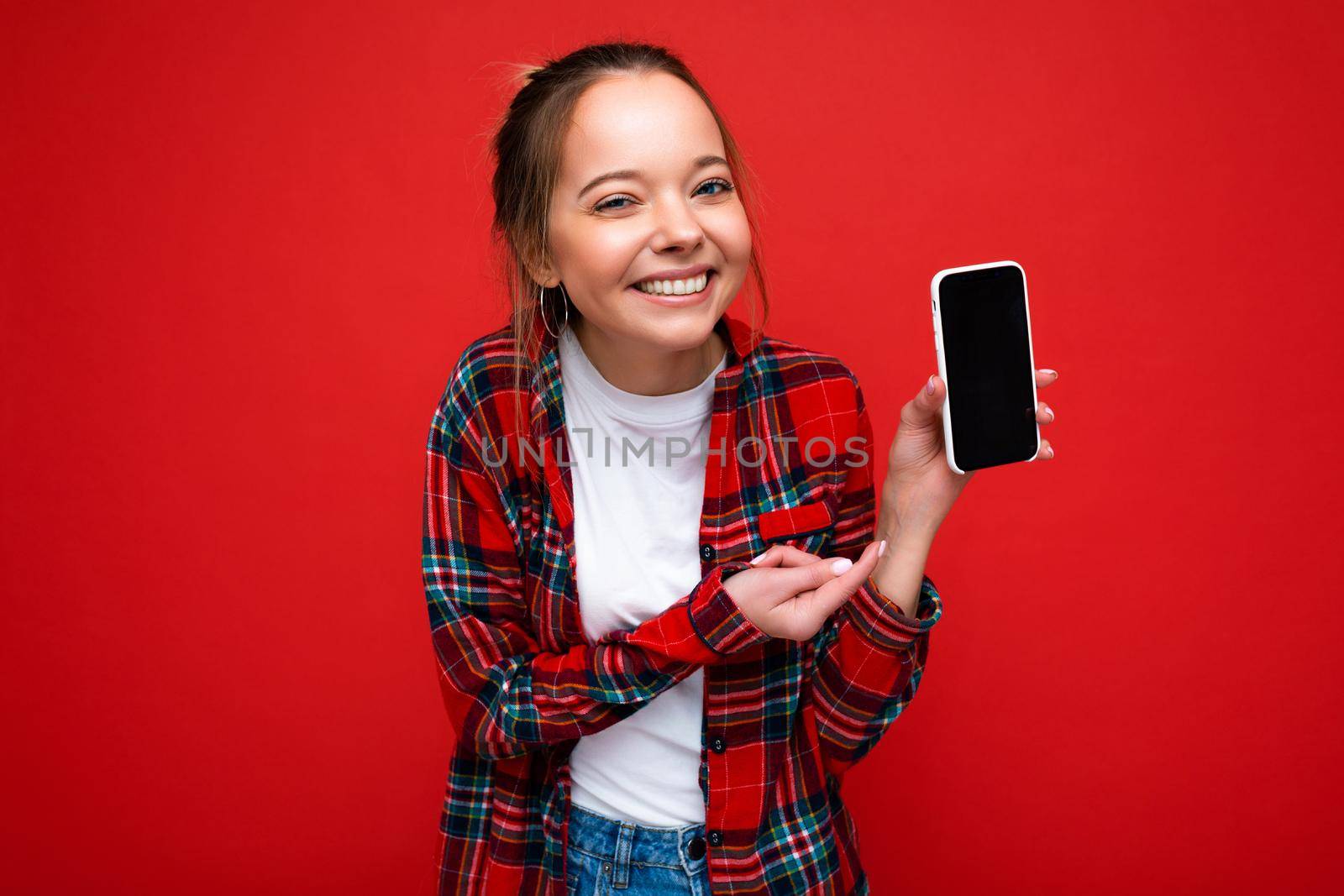 Photo of beautiful smiling young woman good looking wearing casual stylish outfit standing isolated on background with copy space holding smartphone showing phone in hand with empty screen display for mockup pointing at gadjet looking at camera.