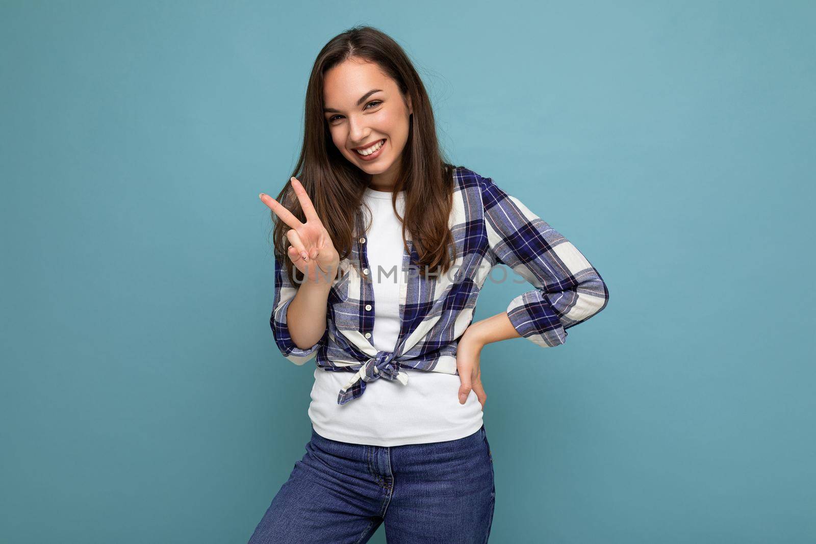 Young positive happy smiling beautiful winsom brunette lady with sincere emotions wearing check shirt poising isolated over blue background with copy space and showing peace gesture.