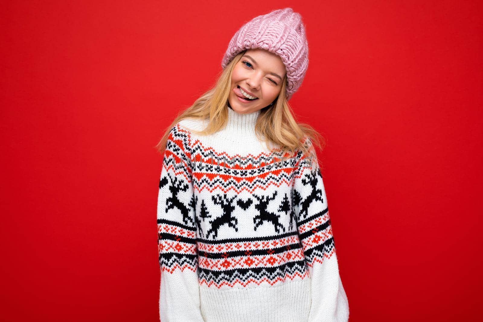 Photo of pretty positive smiling young blonde woman isolated over red background wall wearing winter sweater and pink hat looking at camera and winking.