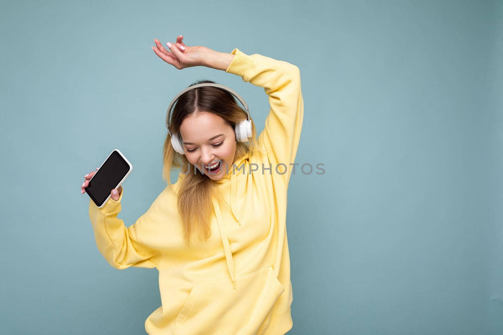 Photo of beautiful joyful smiling young woman wearing stylish casual clothes isolated over background wall holding and using mobile phone wearing white bluetooth headphones listening to music and having fun looking down.