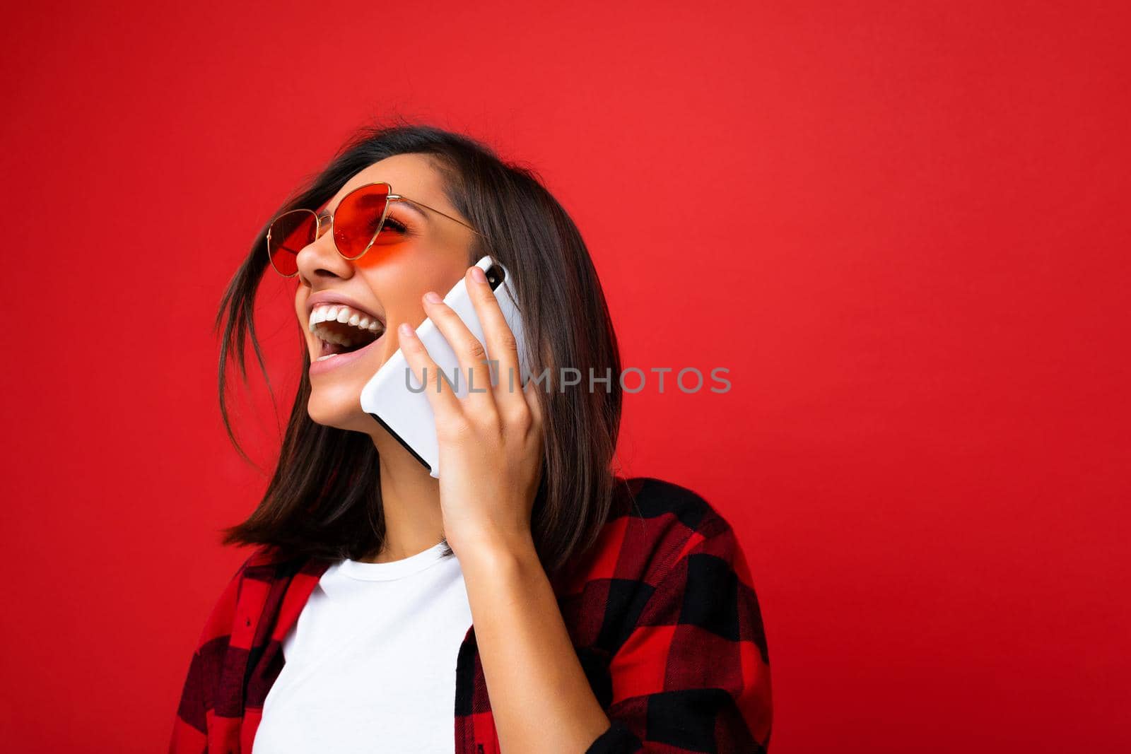 Closeup photo of attractive positive laughing young brunet woman wearing stylish red shirt white t-shirt and red sunglasses isolated over red background communicating on mobile phone looking up.