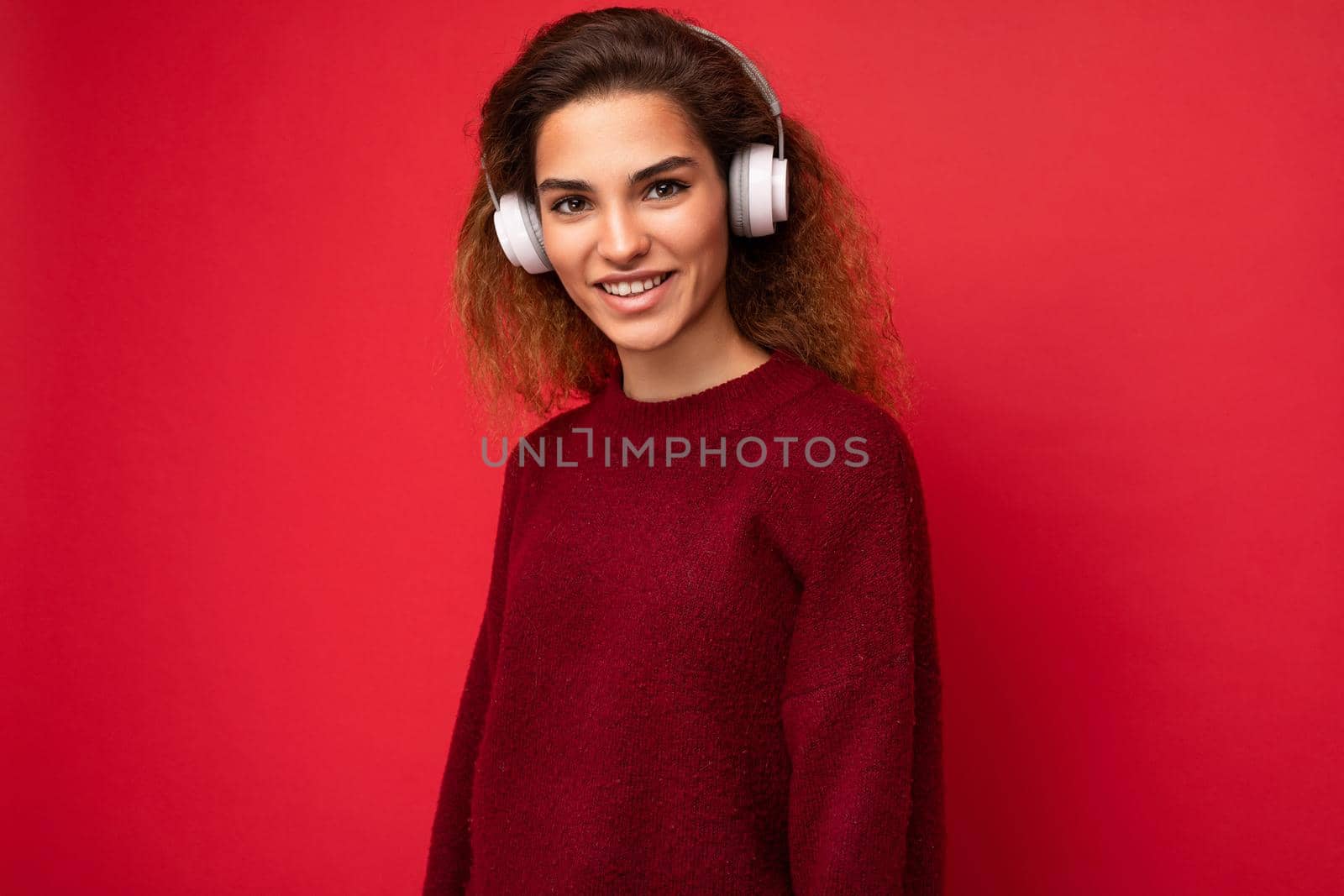 Beautiful happy smiling young brunette curly woman wearing dark red sweater isolated over red background wall wearing white bluetooth headphones listening to music and having fun looking at camera.