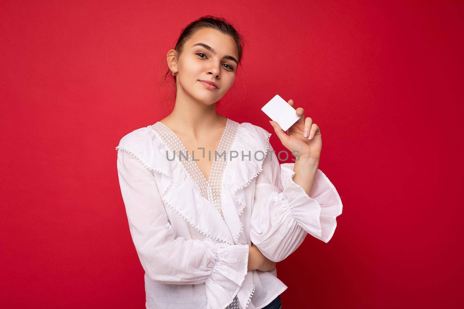 Beautiful happy self-confident young dark blonde woman wearing white blouse isolated over red background holding credit card looking at camera.