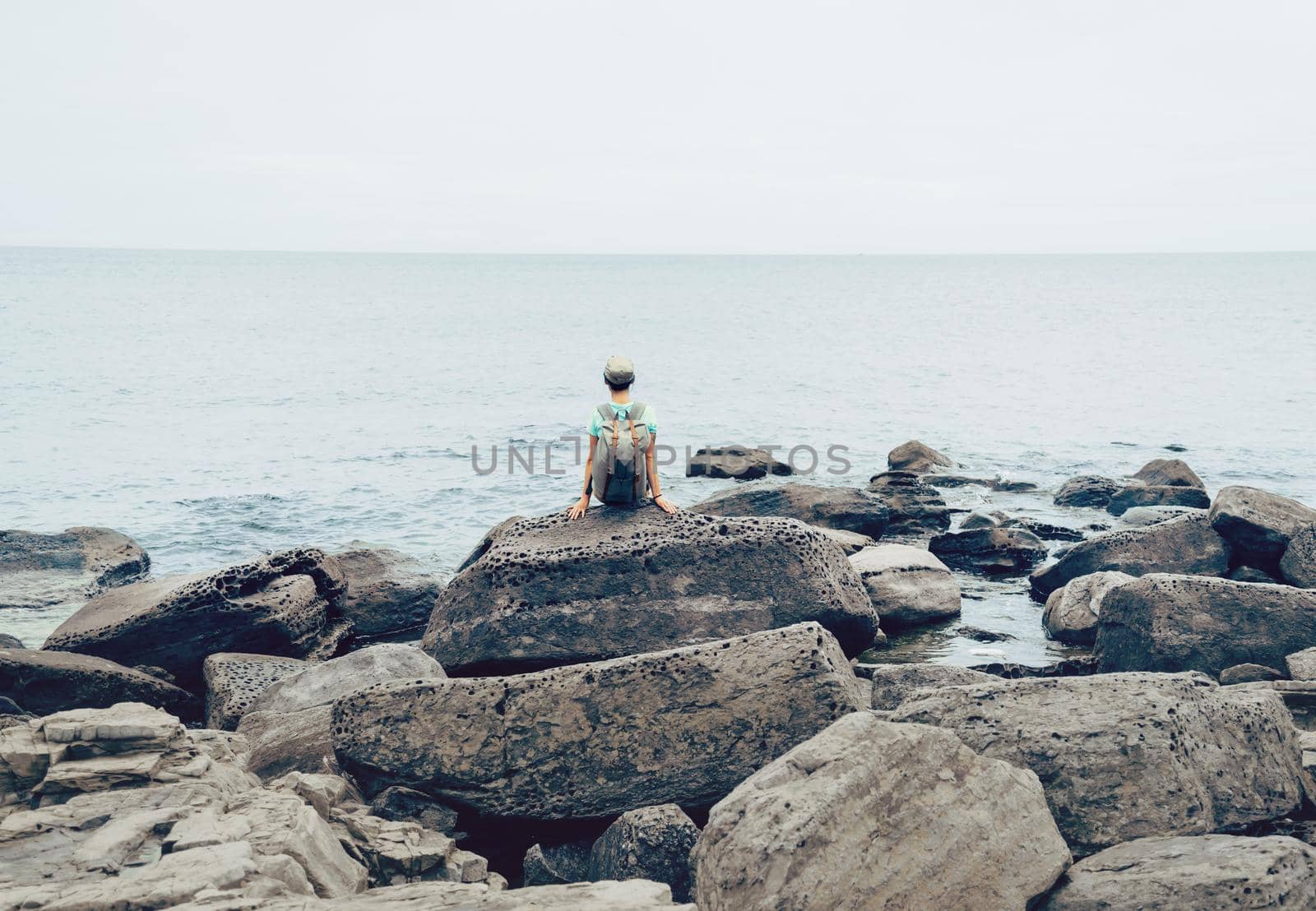 Traveler young woman with backpack resting on big stone near the sea in summer, rear view
