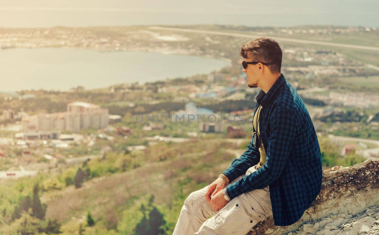 Young man sitting on peak of mountain and looking at town around bay in summer