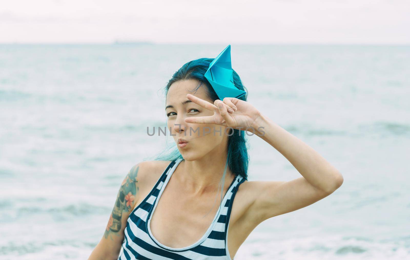 Beautiful young woman with blue hair resting on beach in summer. She raising her arm to her eye with peace sign