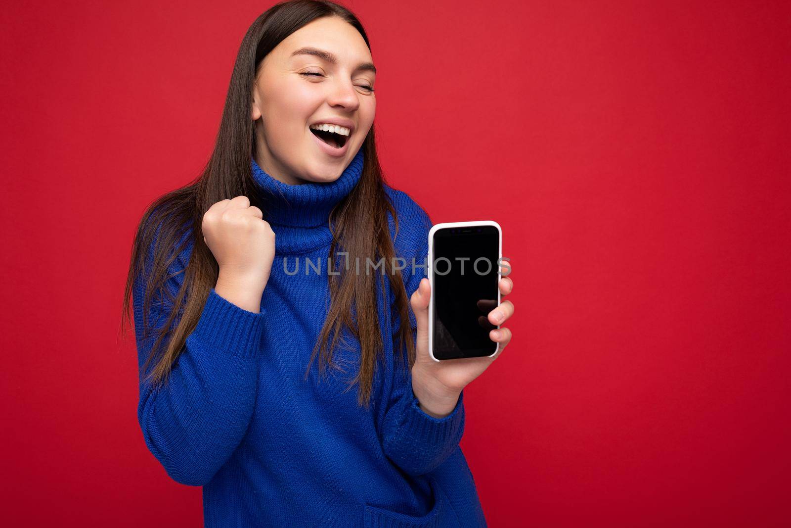 beautiful happy young brunette woman wearing casual blue sweater isolated over red background with empty space holding in hand mobile phone and showing smartphone with empty screen for mockup.