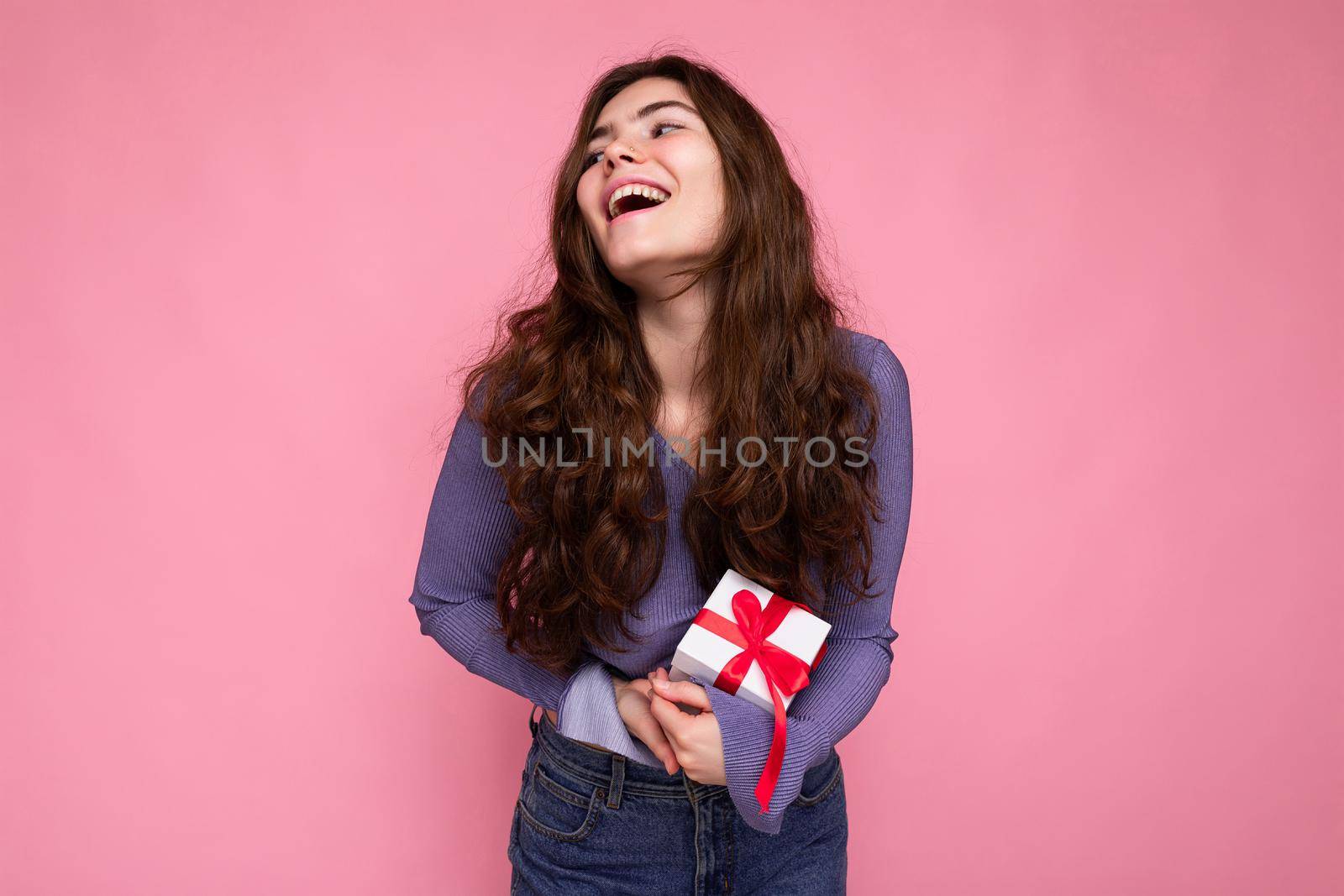 Photo of pretty positive smiling brunette curly young woman isolated over pink background wall wearing purple blouse holding white gift box with red ribbon and having fun.