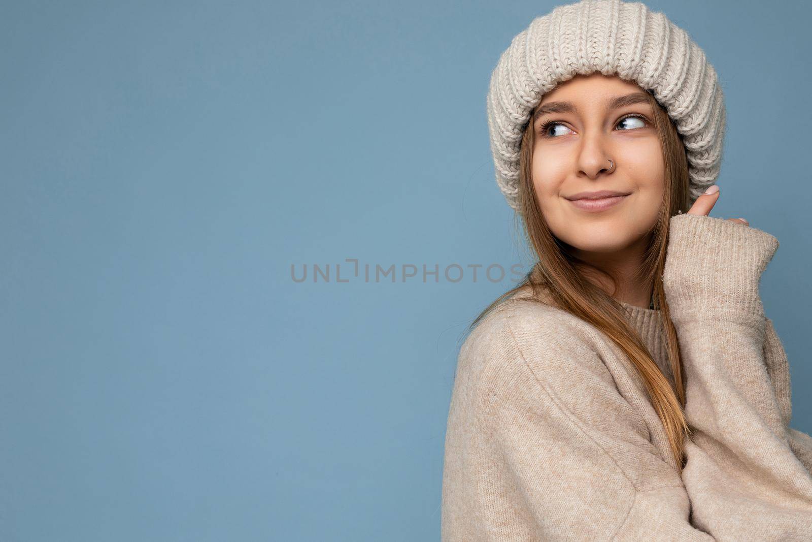 Shot of beautiful sexy smiling adult blonde woman standing isolated on blue background wall wearing beige warm sweater and winter beige hat looking to the side and enjoying. Empty space, copy space