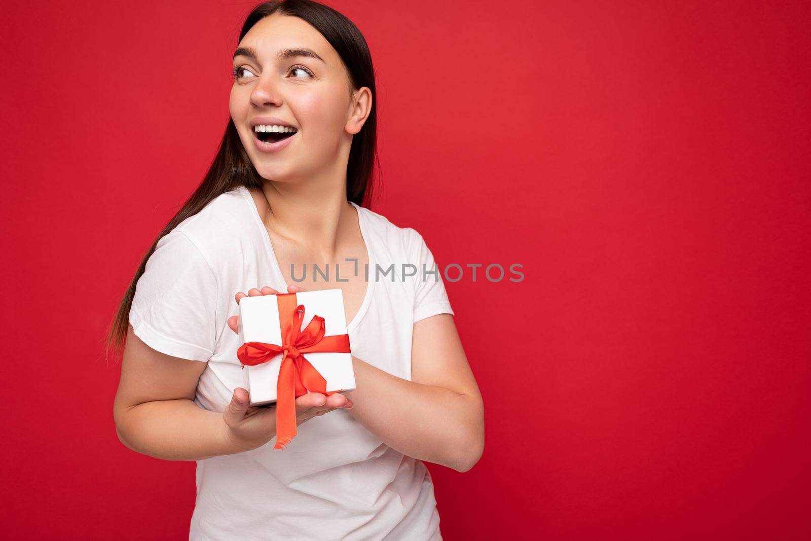 Beautiful happy young brunette woman isolated over colourful background wall wearing stylish casual clothes holding gift box and looking to the side by TRMK