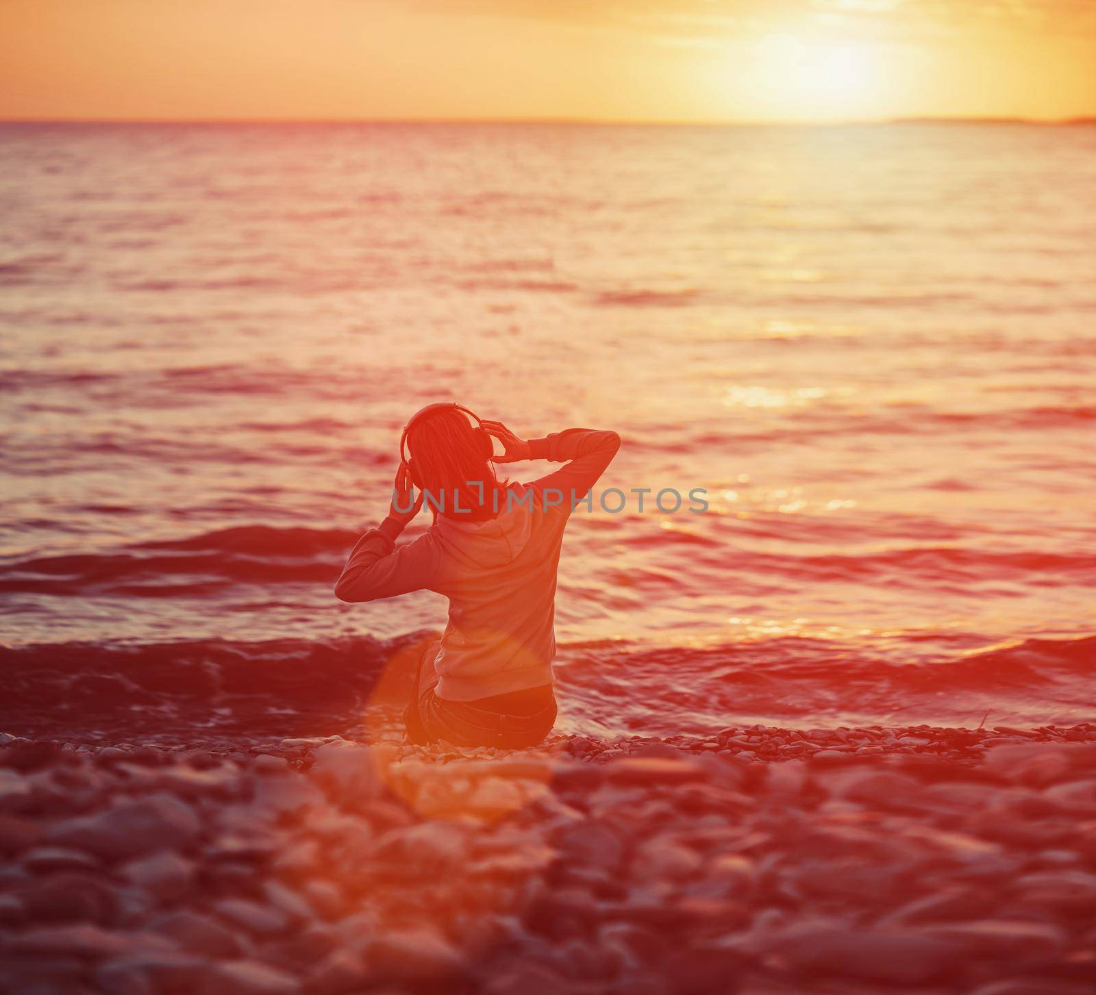 Young woman listening music on beach and enjoying beautiful sunset over the sea. Image with sunlight effect