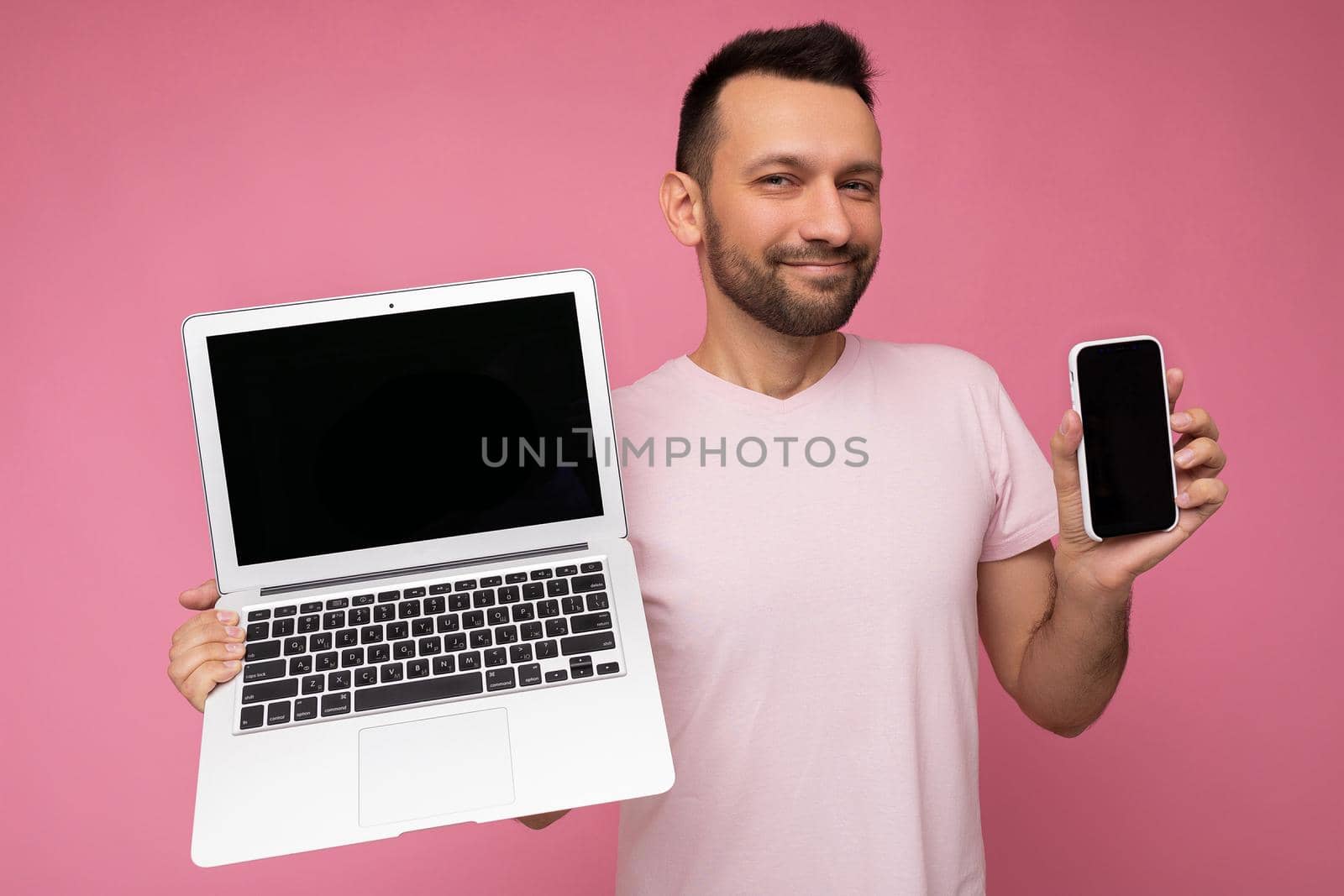 Handsome brunet man holding laptop computer and mobile phone fishily looking at camera in t-shirt on isolated pink background.