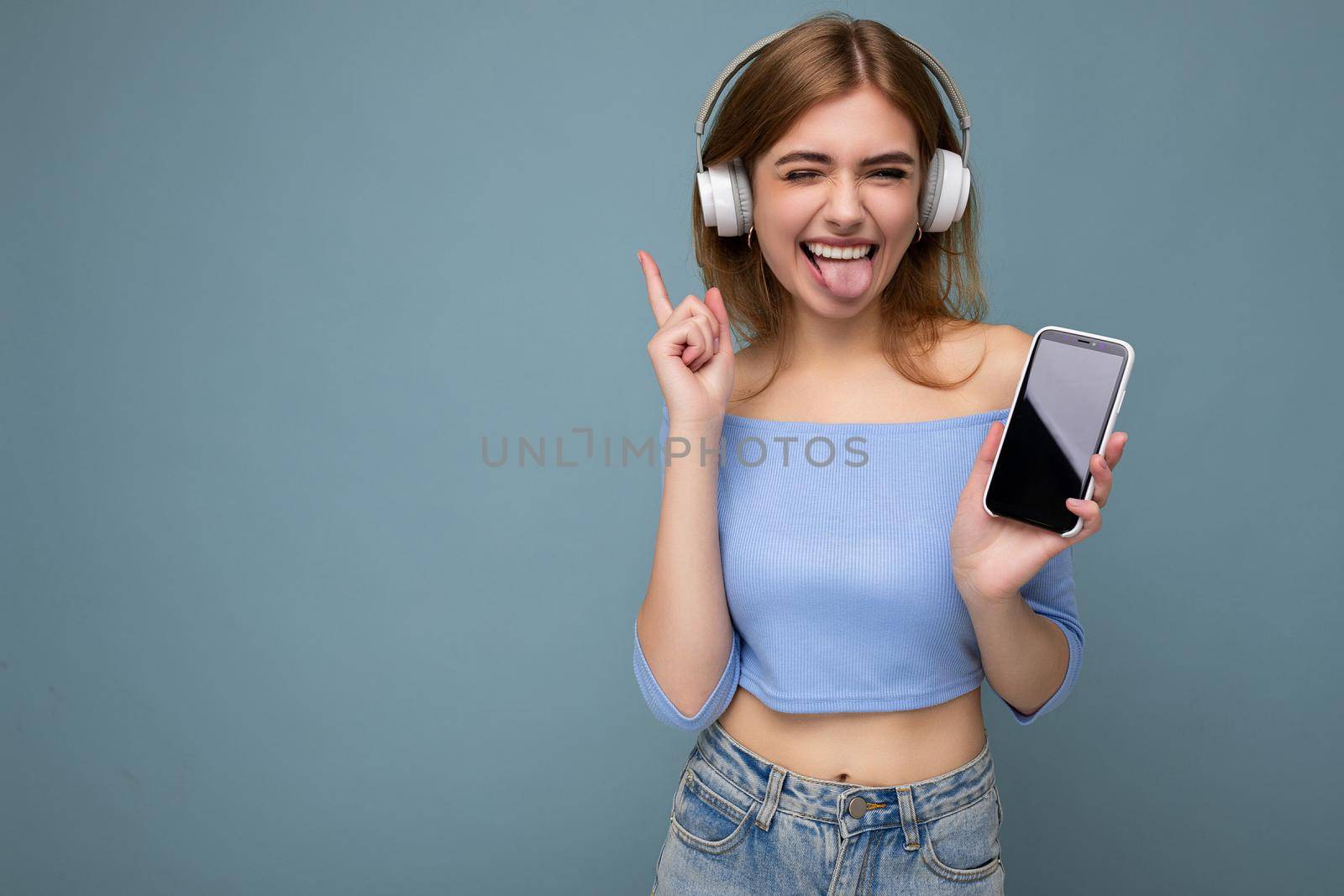 Attractive sexy positive smiling young woman wearing stylish casual outfit isolated on colourful background wall holding and showing mobile phone with empty screen for cutout wearing white bluetooth headphones and having fun looking at camera. copy space