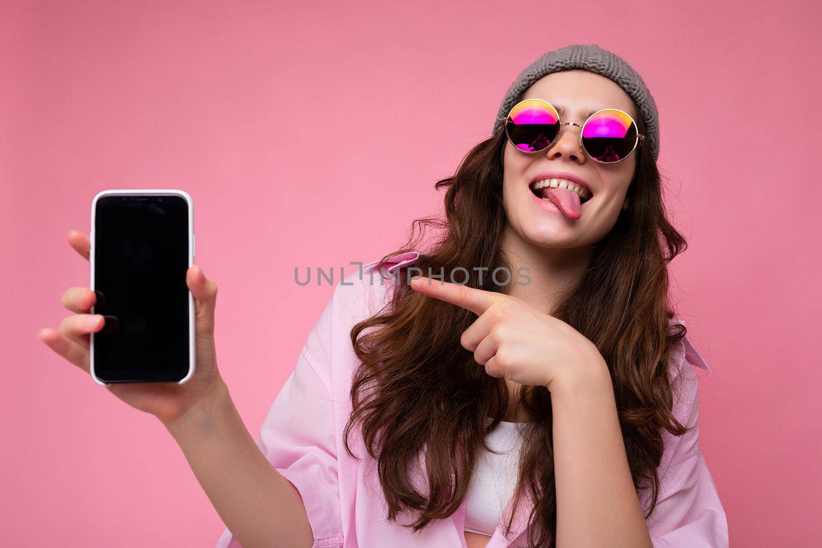 Positive attractive young brunette woman wearing stylish pink shirt grey hat and colourful sunglasses isolated over pink background holding in hand and showing mobile phone with empty display for mockup looking at camera pointing finger at gadjet and showing tongue by TRMK