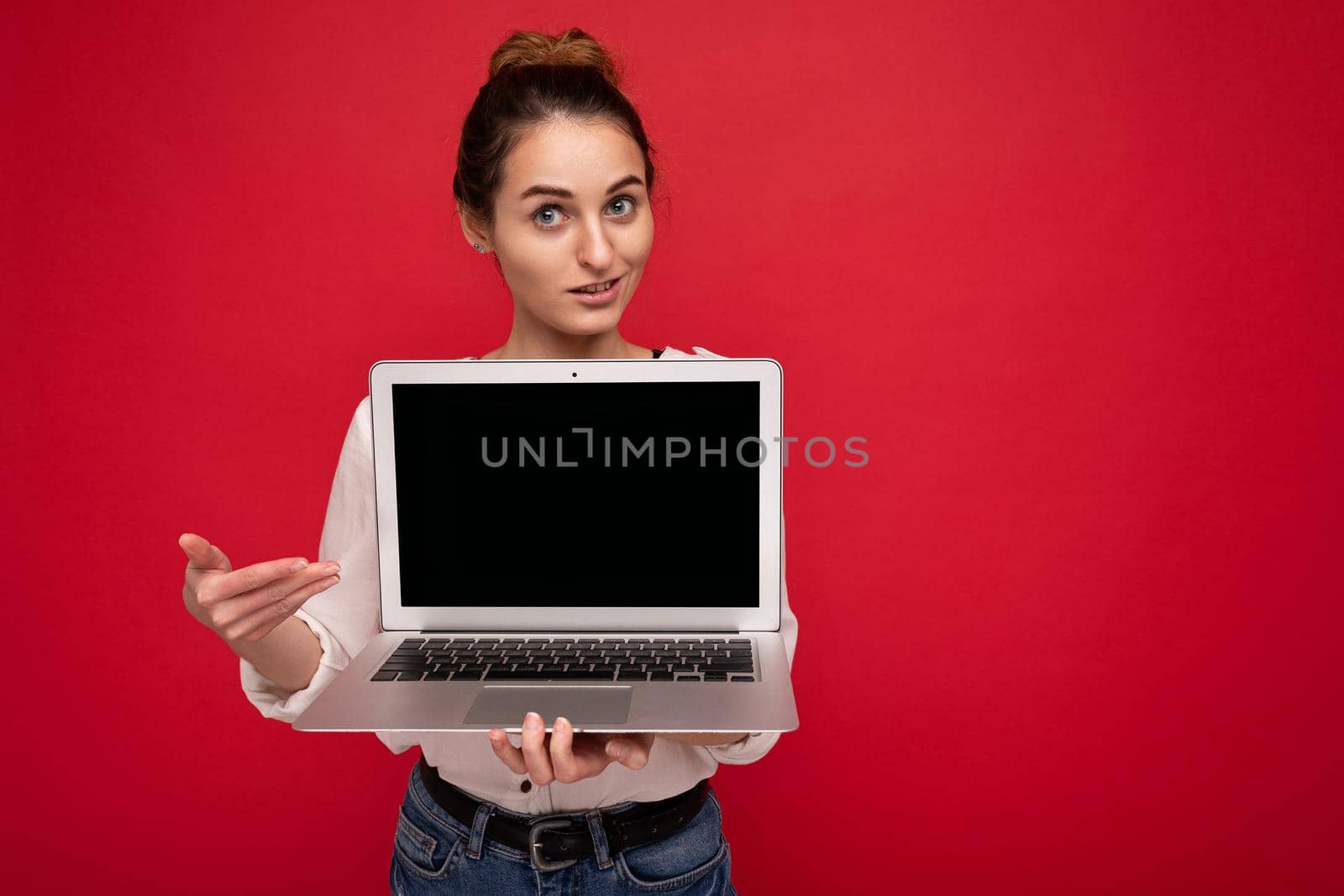Close-up portrait of Beautiful smiling happy young woman holding computer laptop looking at camera pointing at display wearing casual smart clothes isolated over red wall background.