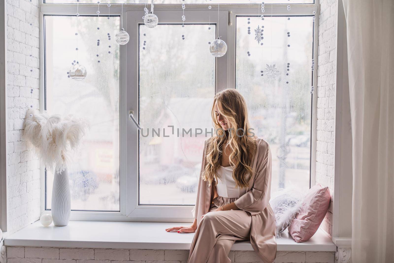 Resting and thinking woman in pajamas sitting on the window-sill at home. Side view. by Matiunina