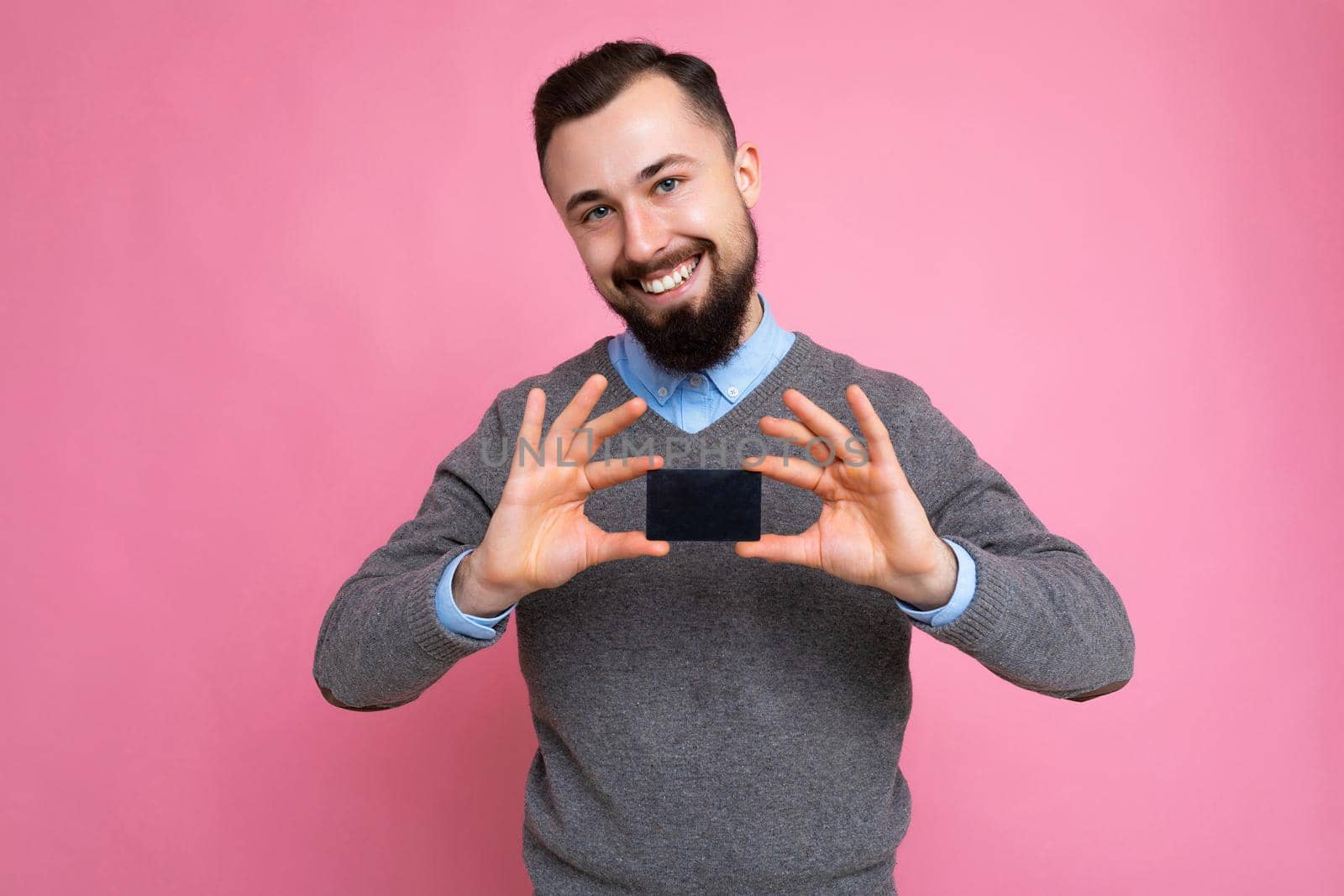 Handsome smiling brunette bearded man wearing grey sweater and blue shirt isolated on background wall holding credit card looking at camera.
