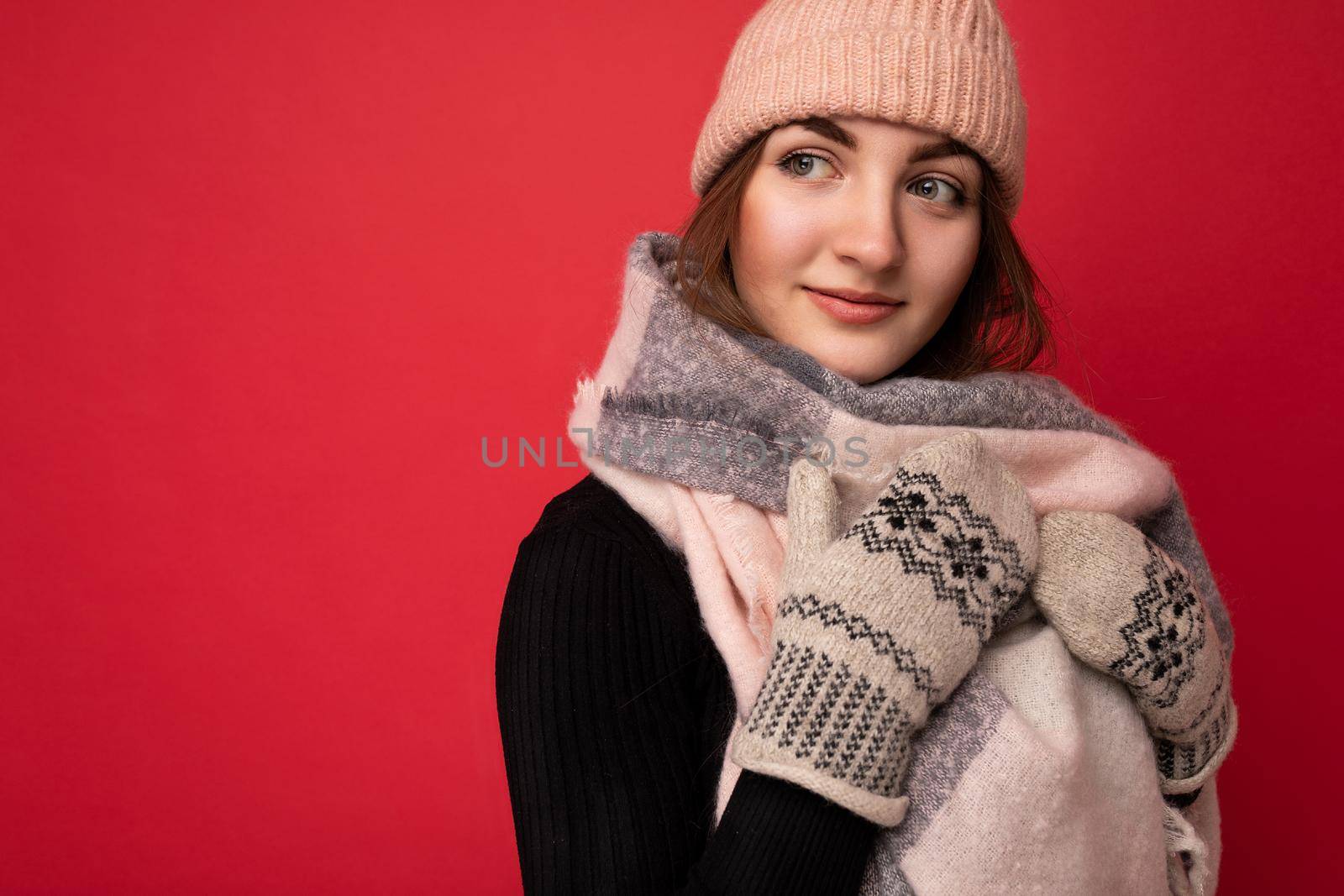 Closeup photo of beautiful happy cute young brunette woman isolated over red background wall wearing winter scarf mittens and warm hat looking to the side.