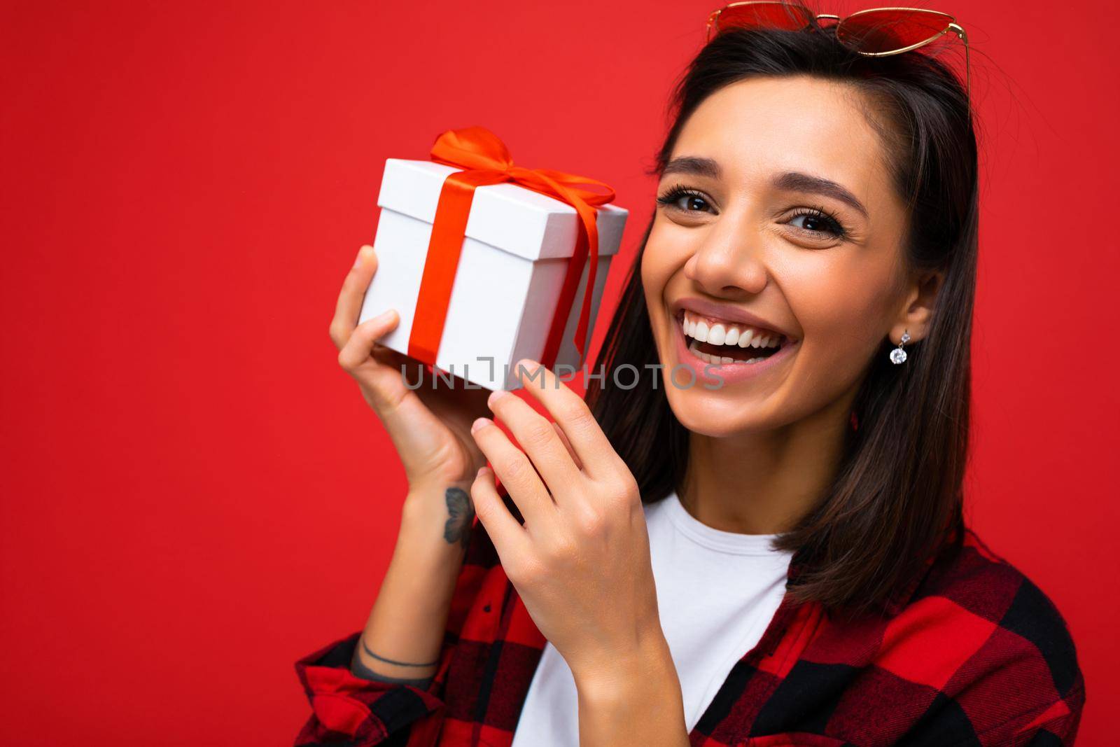 Shot of attractive positive smiling young brunette woman isolated over colourful background wall wearing everyday trendy outfit holding gift box and looking at camera.