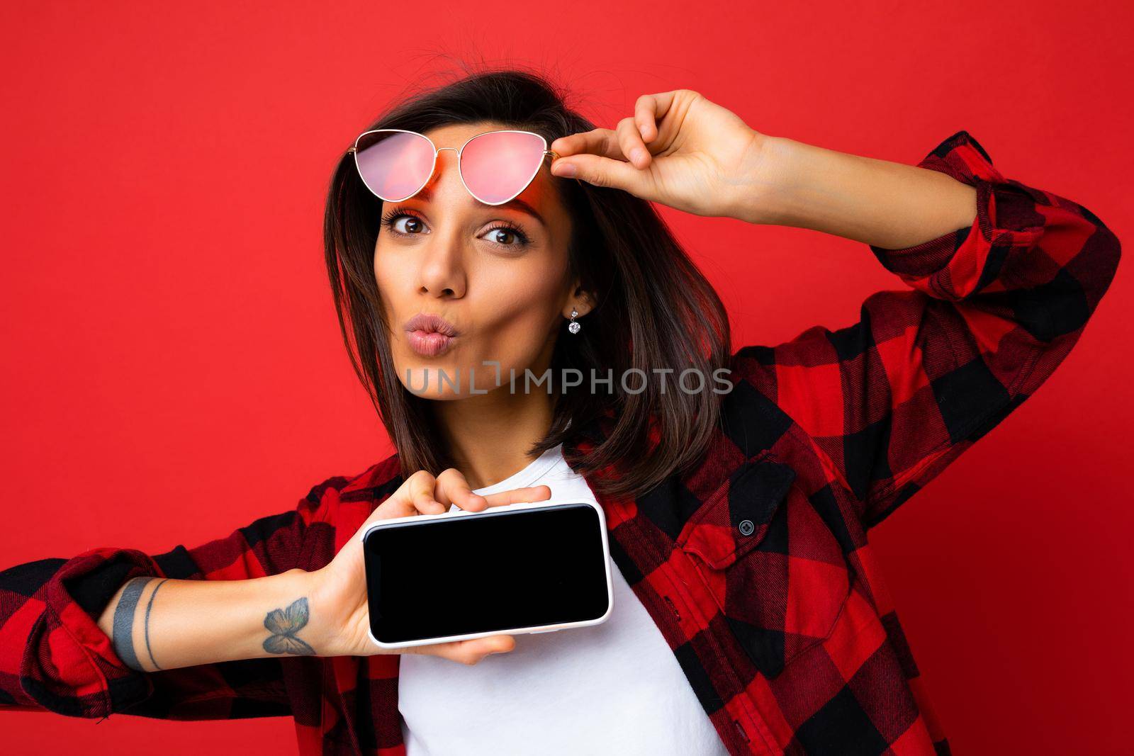 Photo of beautiful smiling young woman good looking wearing casual stylish red shirt white t-shirt and red sunglasses standing isolated on red background with copy space holding smartphone showing phone in hand with empty screen display for mockup looking at camera and giving kiss by TRMK
