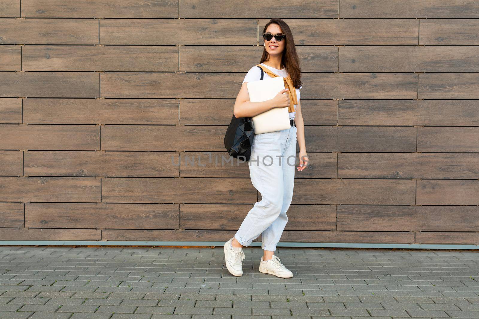 Full length photo shot of young charming beautifyl brunet woman going in the street near brown wall looking to side in white t-shirt and light blue jeans white sneakers with black female bag holding computer laptop and listening to music with wireless headphones.