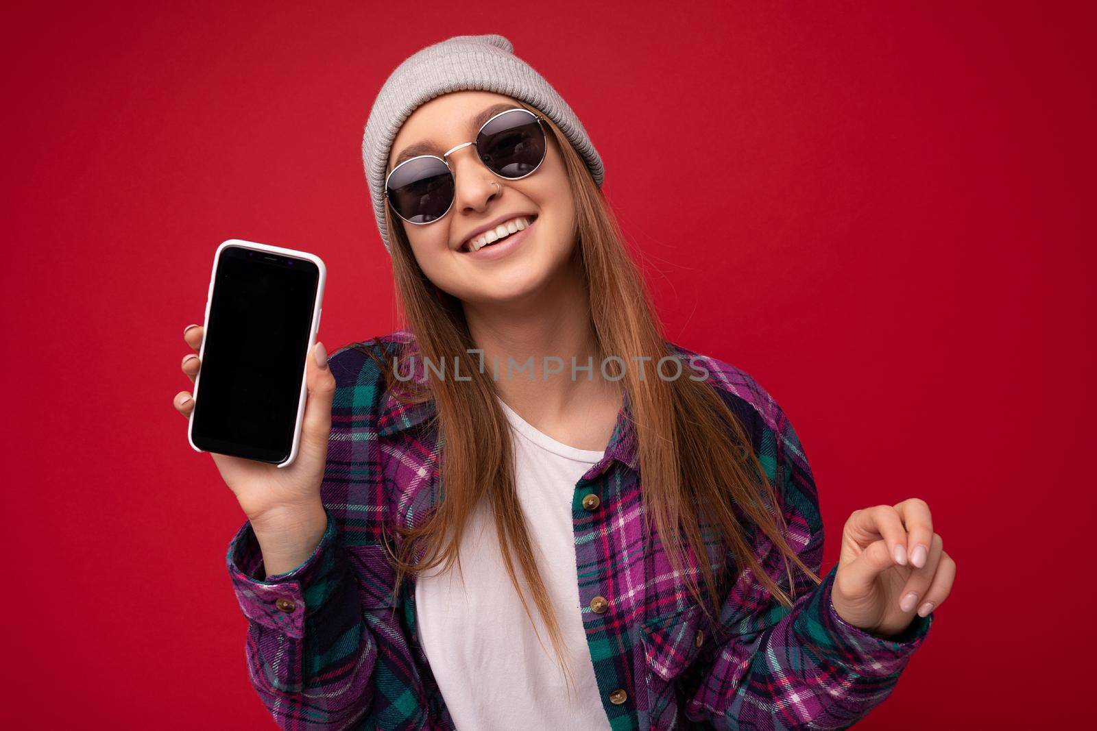 Closeup photo of attractive smiling positive young blonde woman wearing stylish purple shirt and casual white t-shirt grey hat and sunglasses isolated over red background holding in hand and showing mobile phone with empty screen for mockup looking at camera by TRMK