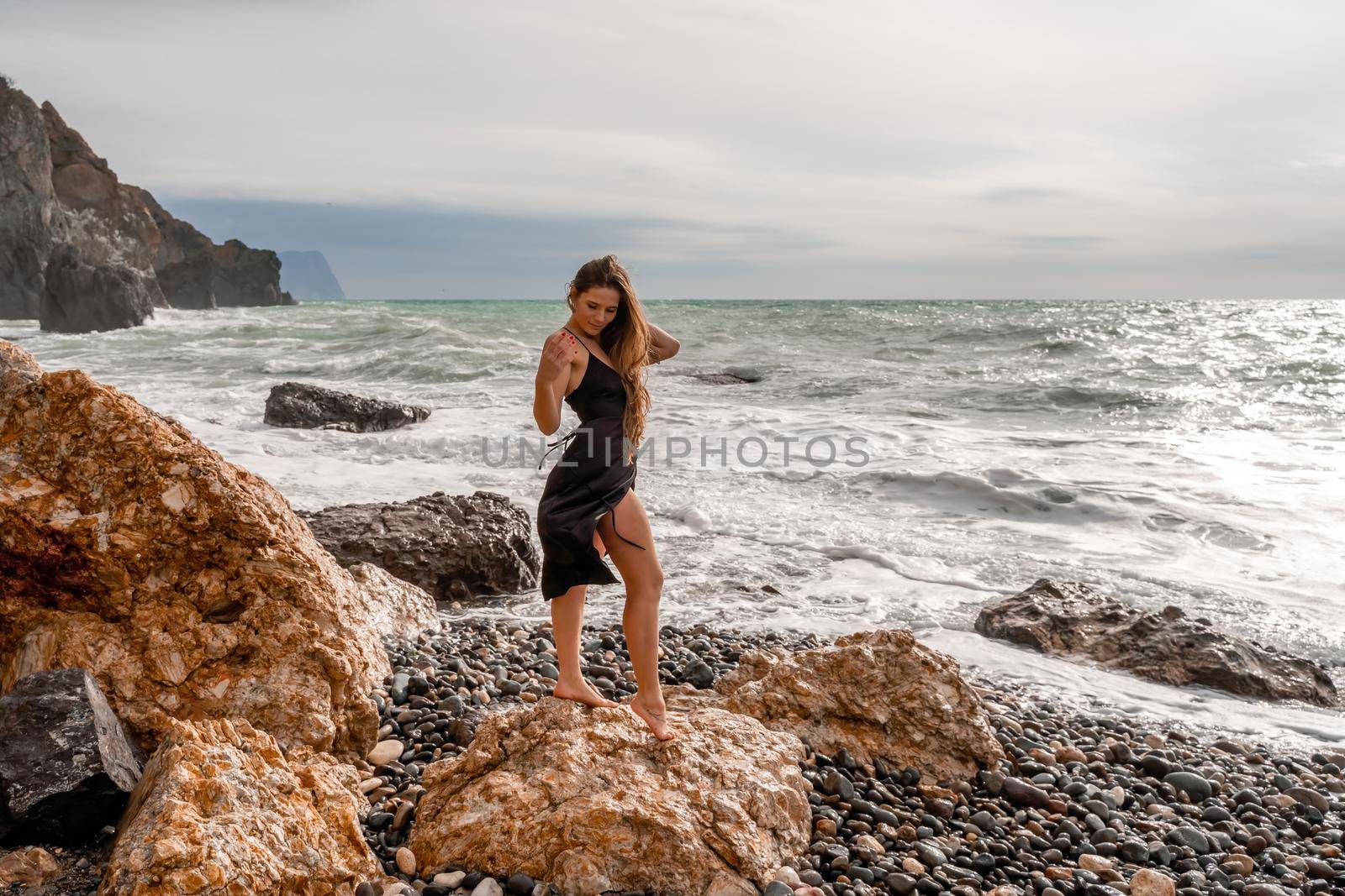 A beautiful girl in a black dress stands on a rock, big waves with white foam. A cloudy stormy day at sea, with clouds and big waves hitting the rocks. by Matiunina