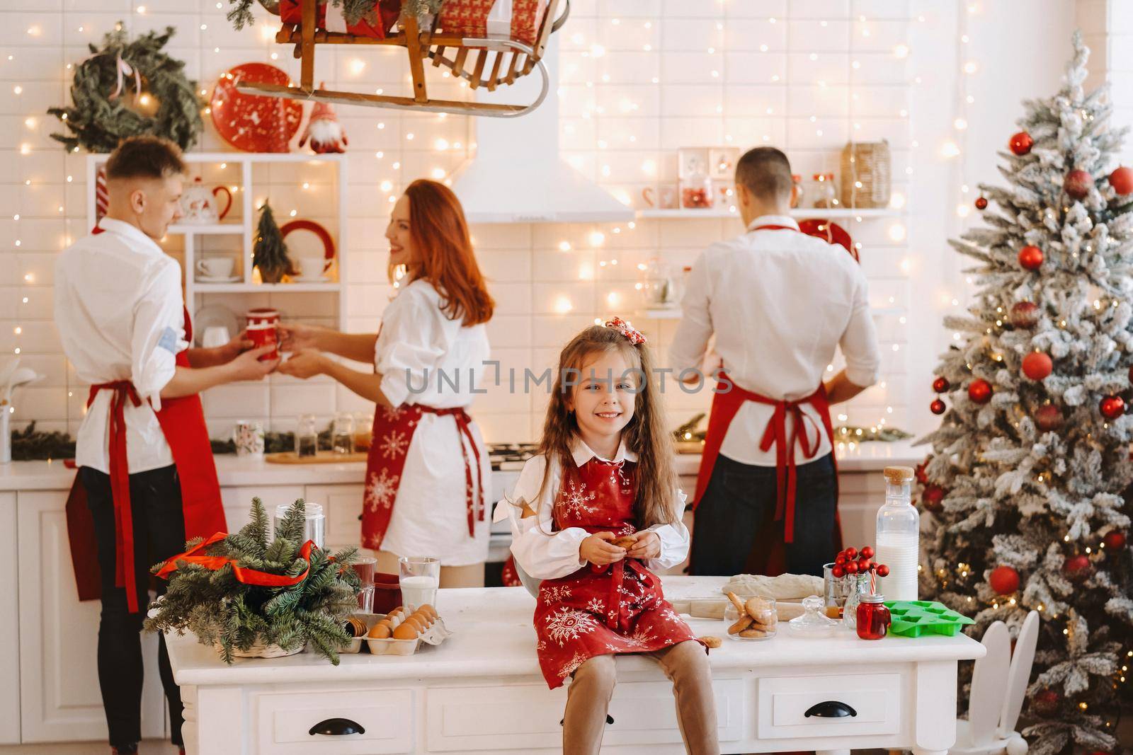 A little girl in the New Year's kitchen is sitting on the table with cookies in her hands and her parents are preparing Christmas dinner by Lobachad