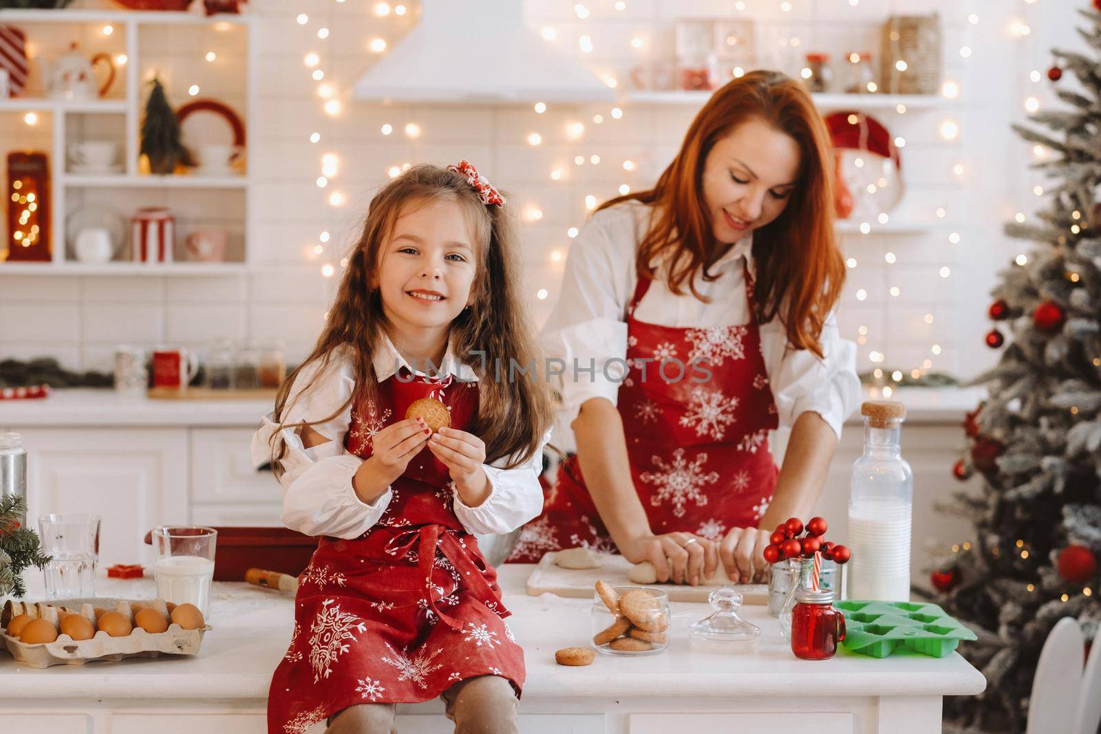 A little girl in the New Year's kitchen is sitting on the table with cookies in her hands, and her mother is preparing dough.