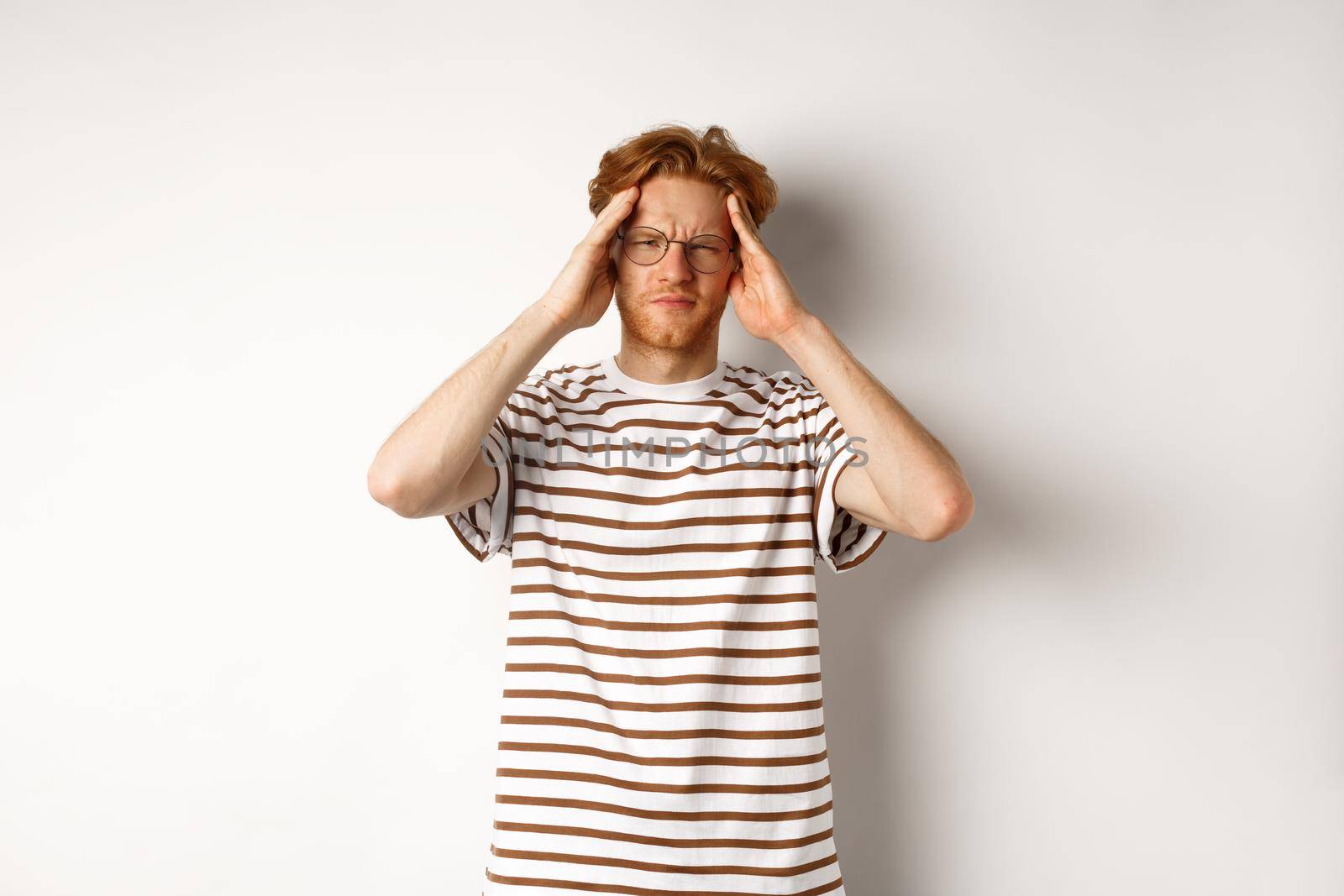 Image of young man with red hair and glasses touching head, frowning from painful migraine, having headache, standing over white background.