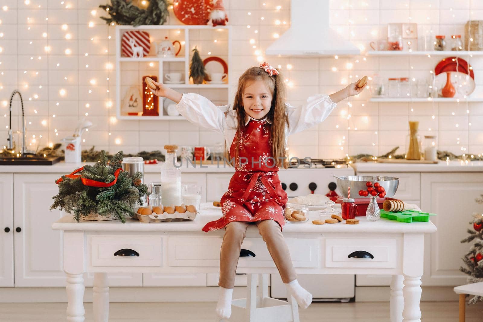 A happy little girl in the Christmas kitchen is sitting on the table with cookies in her hands by Lobachad