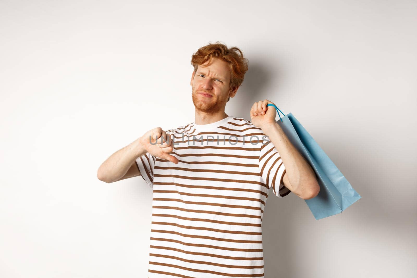 Disappointed young man with red hair and beard showing thumbs-down after bad shopping experience, holding bag over shoulder and frowning upset, white background.
