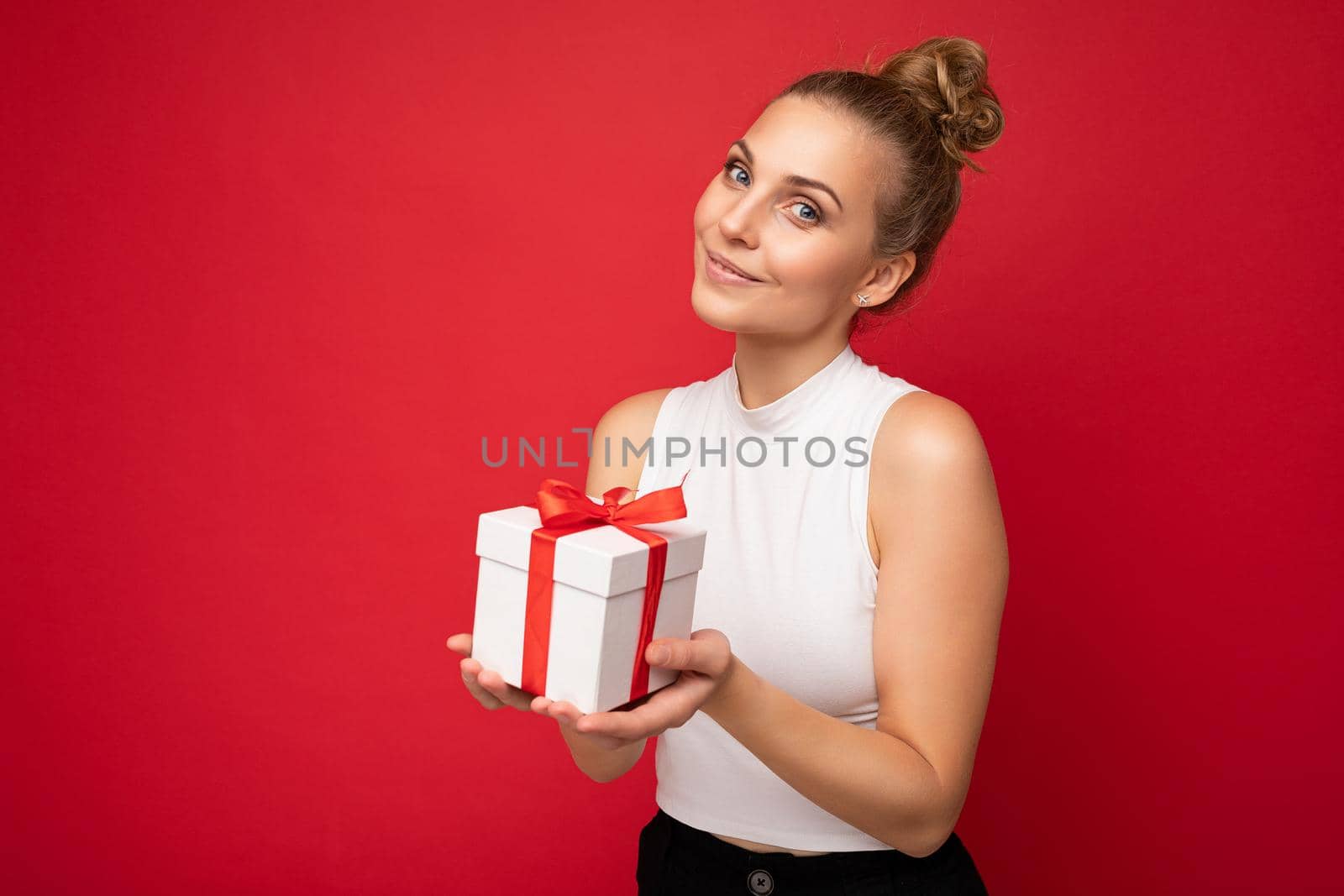 Pretty happy young blonde female person isolated over red background wall wearing white top holding gift box and looking at camera by TRMK