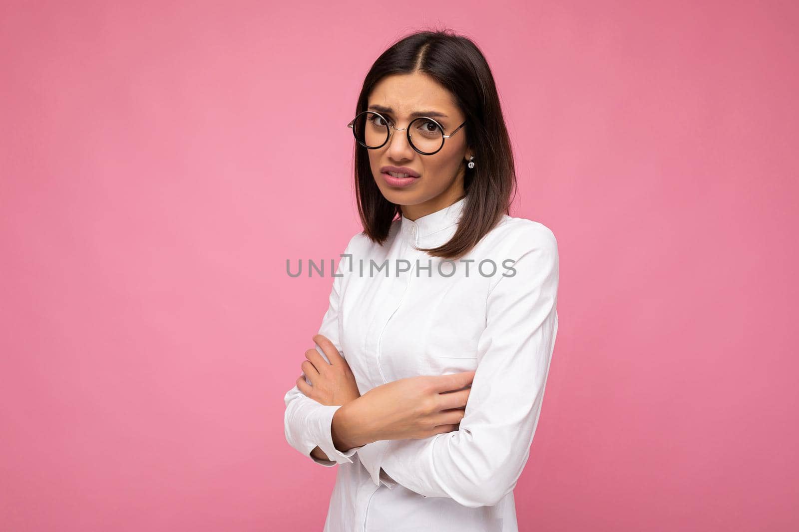 Shot of pretty dissatisfied angry offended young brunette woman wearing white shirt and stylish optical glasses isolated over pink background looking at camera by TRMK