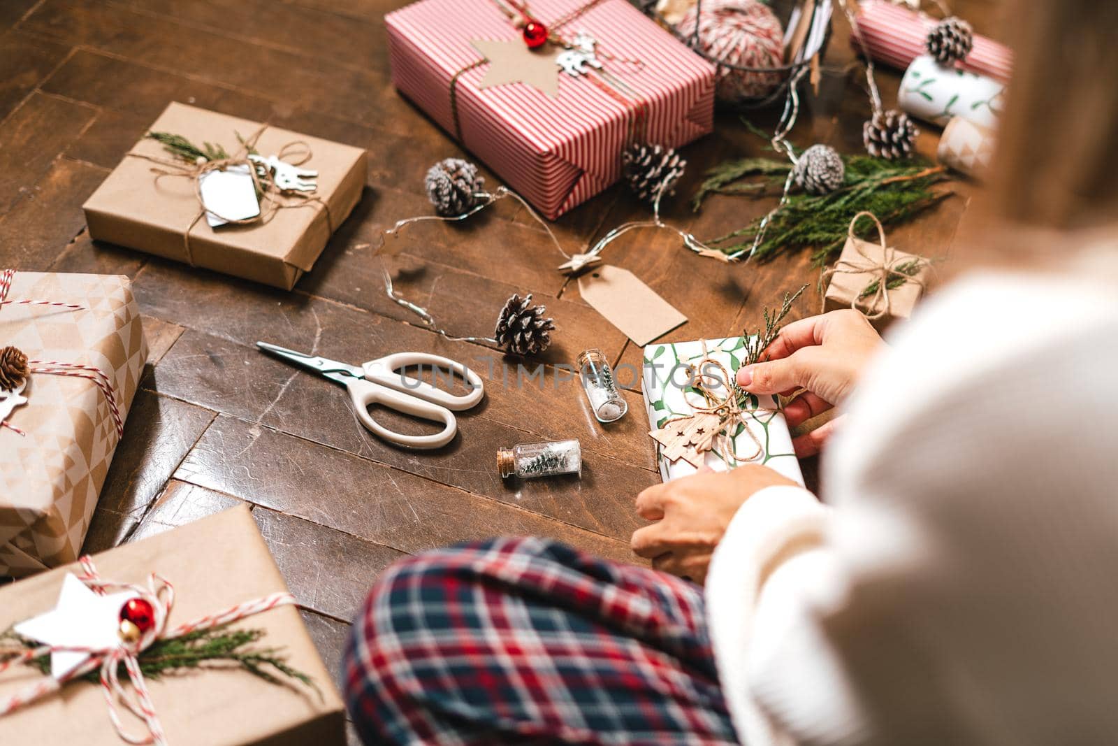 Woman s hands wrapping Christmas gift boxes, close up. Cropped female sit and preparing natural eco presents on floor with decor elements and items. Merry Christmas or New year DIY packing Concept.