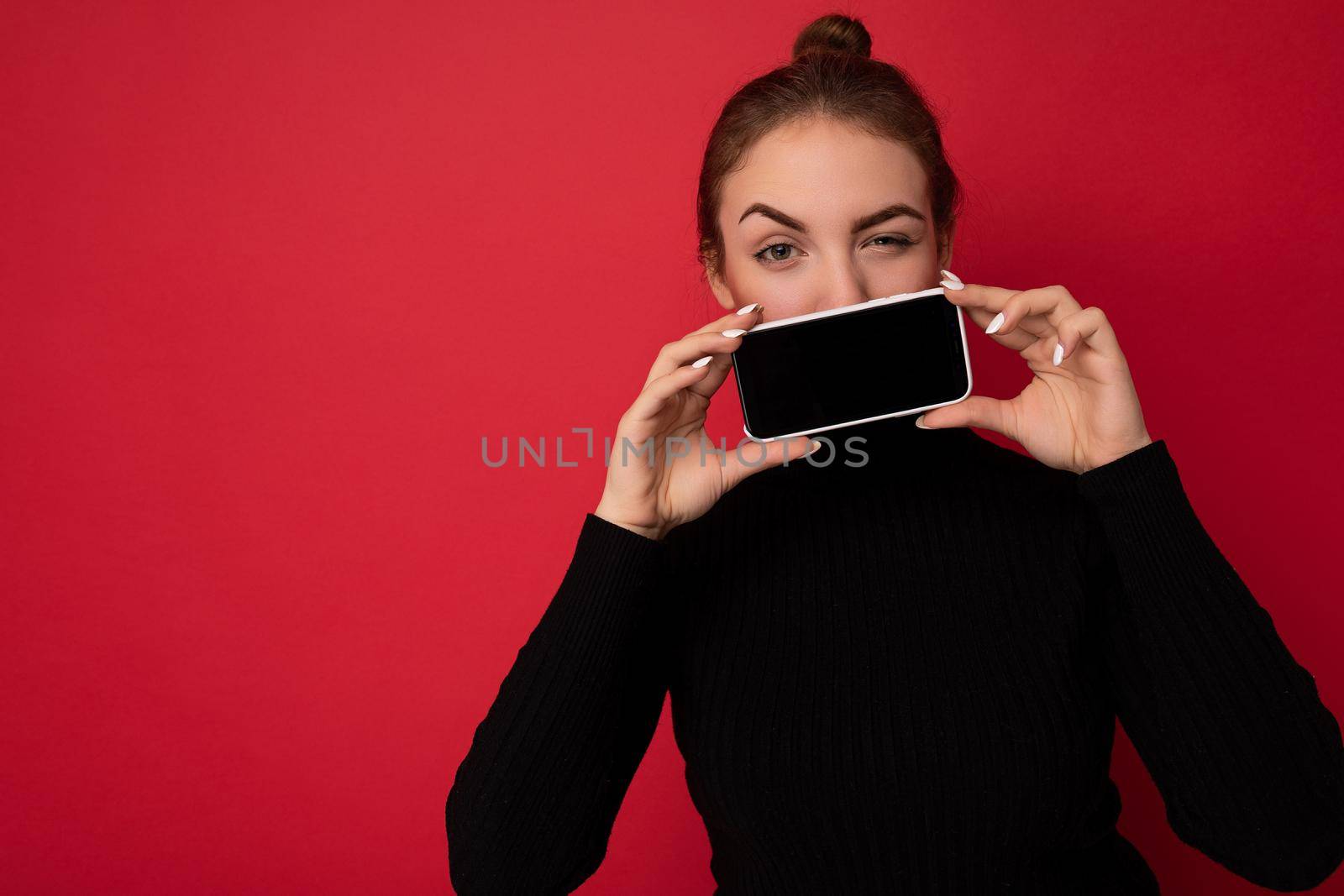 Photo of attractive winking young brunette woman wearing black sweater standing isolated over red background showing mobile phone with empty screen for mockup looking at camera by TRMK